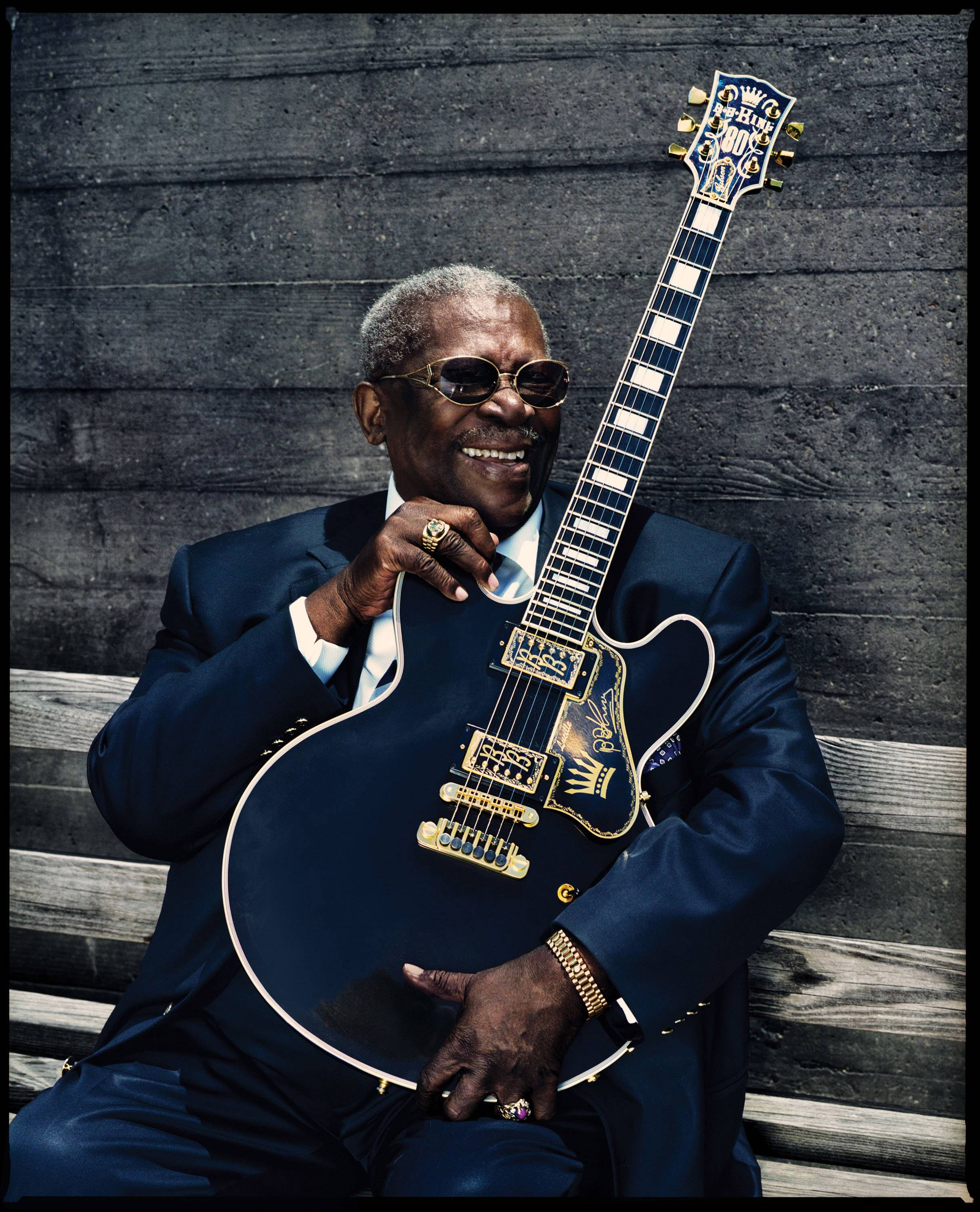 藍調傳奇B.B. King 壁紙 — 一位年長的黑人男性，身穿剪裁俐落的藍色西裝、戴著太陽眼鏡，對著一面風化的木牆，手持一把訂製的藍色電結他，笑容滿面。