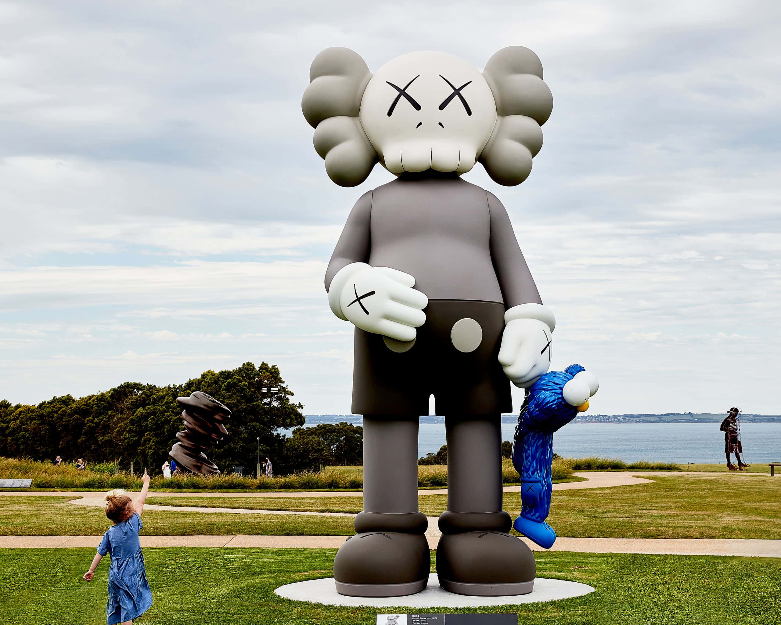 藝術家 KAWS 的經典作品展現視覺魅力 壁紙 — 一個巨大的灰色KAWS雕塑，帶有X形眼睛，站在水邊的草地上，一個穿藍色衣服的孩子正伸手觸碰它。