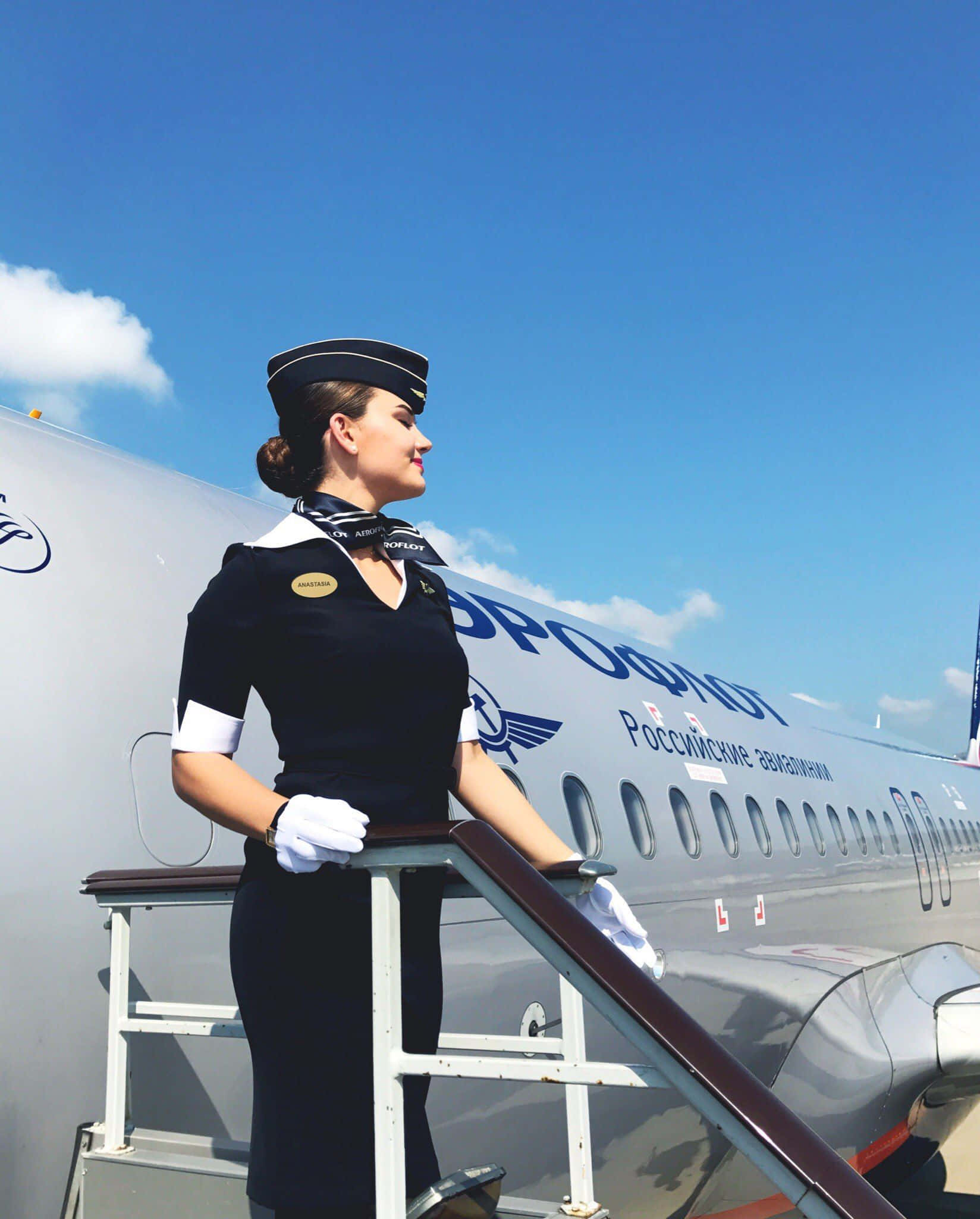 藝術風格的空服員照片 壁紙 — 一位優雅的女空中服務員穿著深色制服，在晴朗的藍天下優雅地站在俄羅斯航空公司的飛機樓梯上。