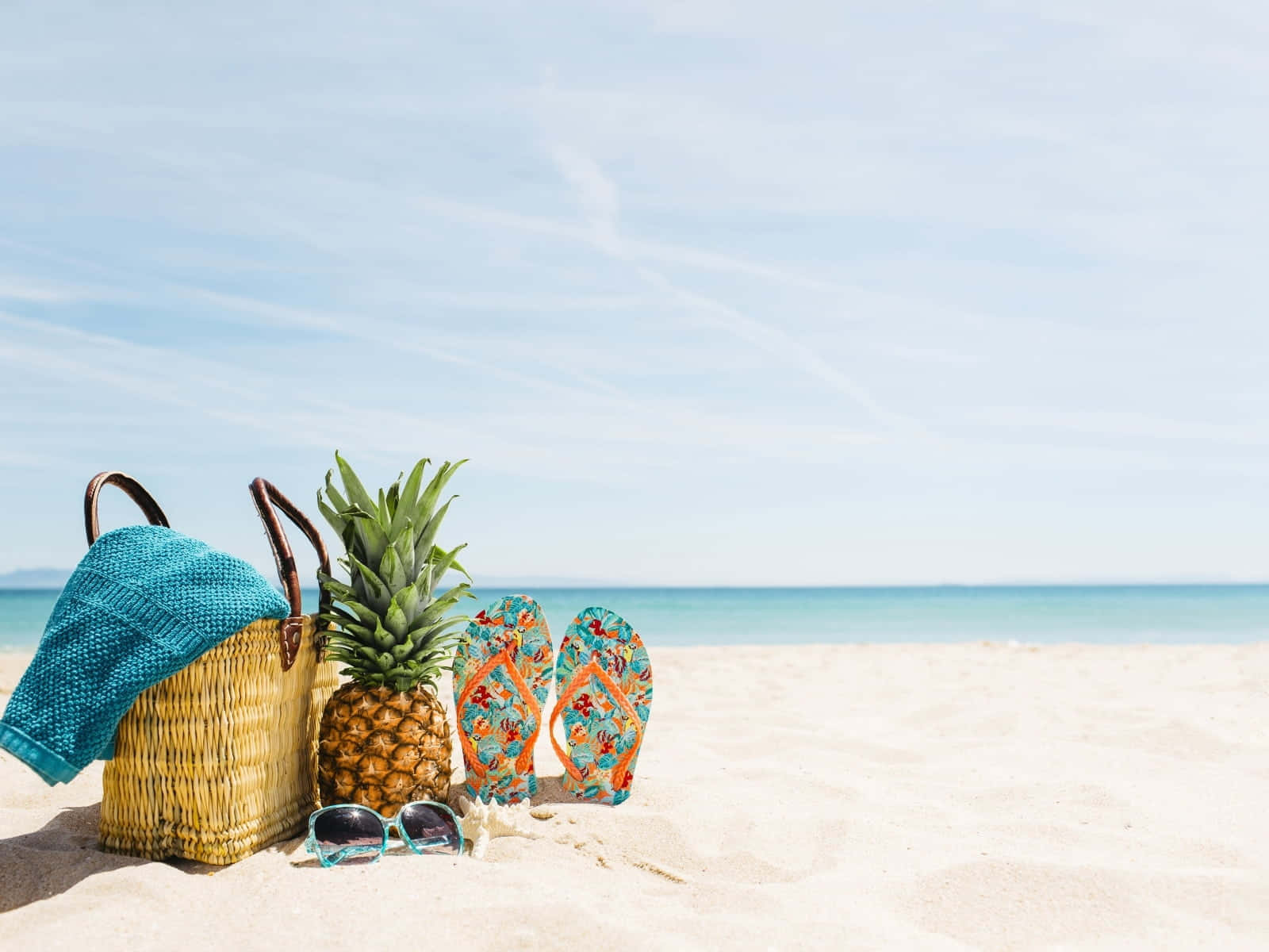 Beach Bag 桌布 壁紙 — 藤織沙灘袋、藍綠色毛巾、菠蘿人字拖、太陽眼鏡和防曬霜放在陽光普照的沙灘上，旁邊是平靜的藍綠色海洋，天空清澈湛藍。
