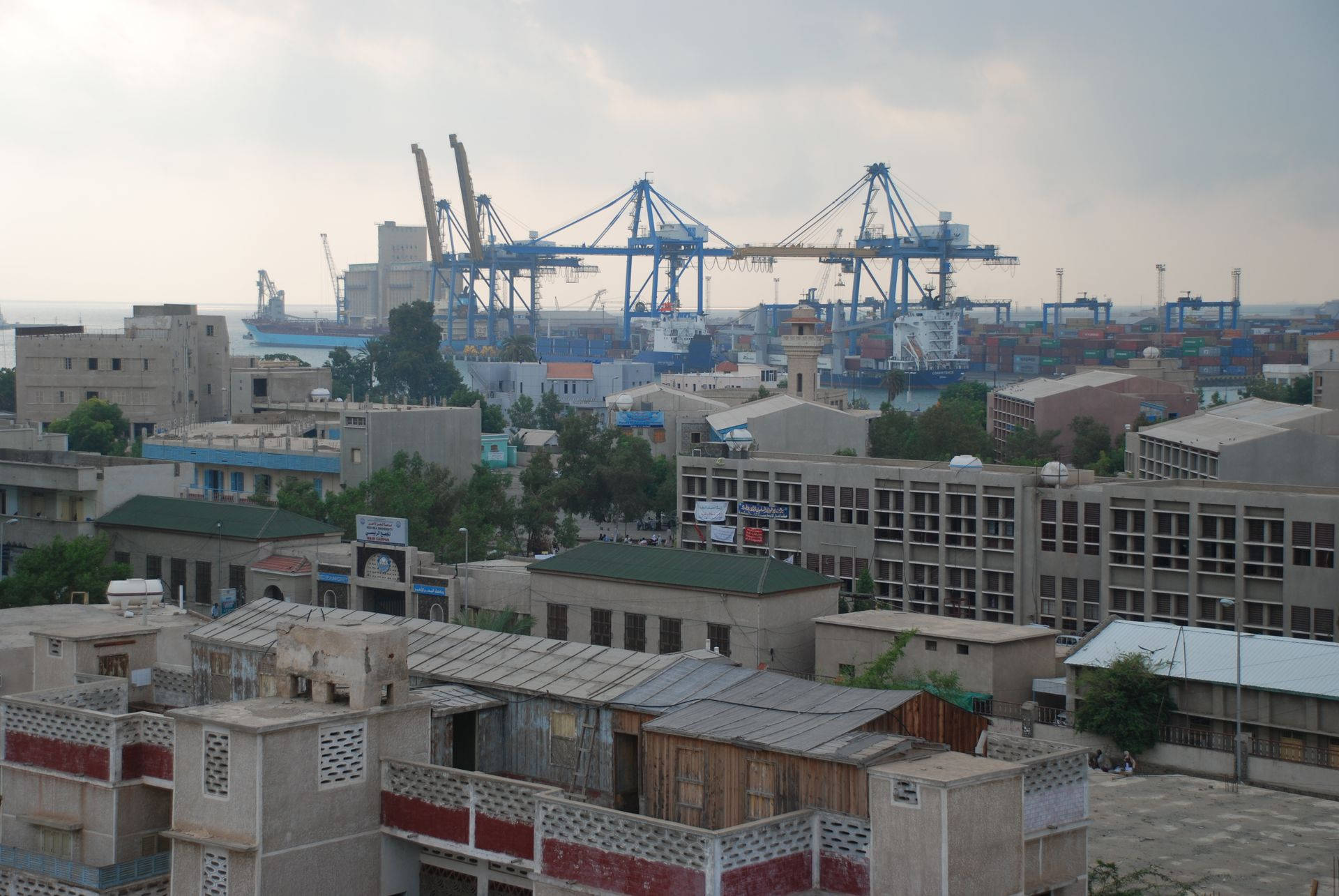 蘇丹住宅區 壁紙 — 廣闊的城市景觀，前景是風化的建築物，背景是巨大的藍色港口起重機，在陰天的主宰下。