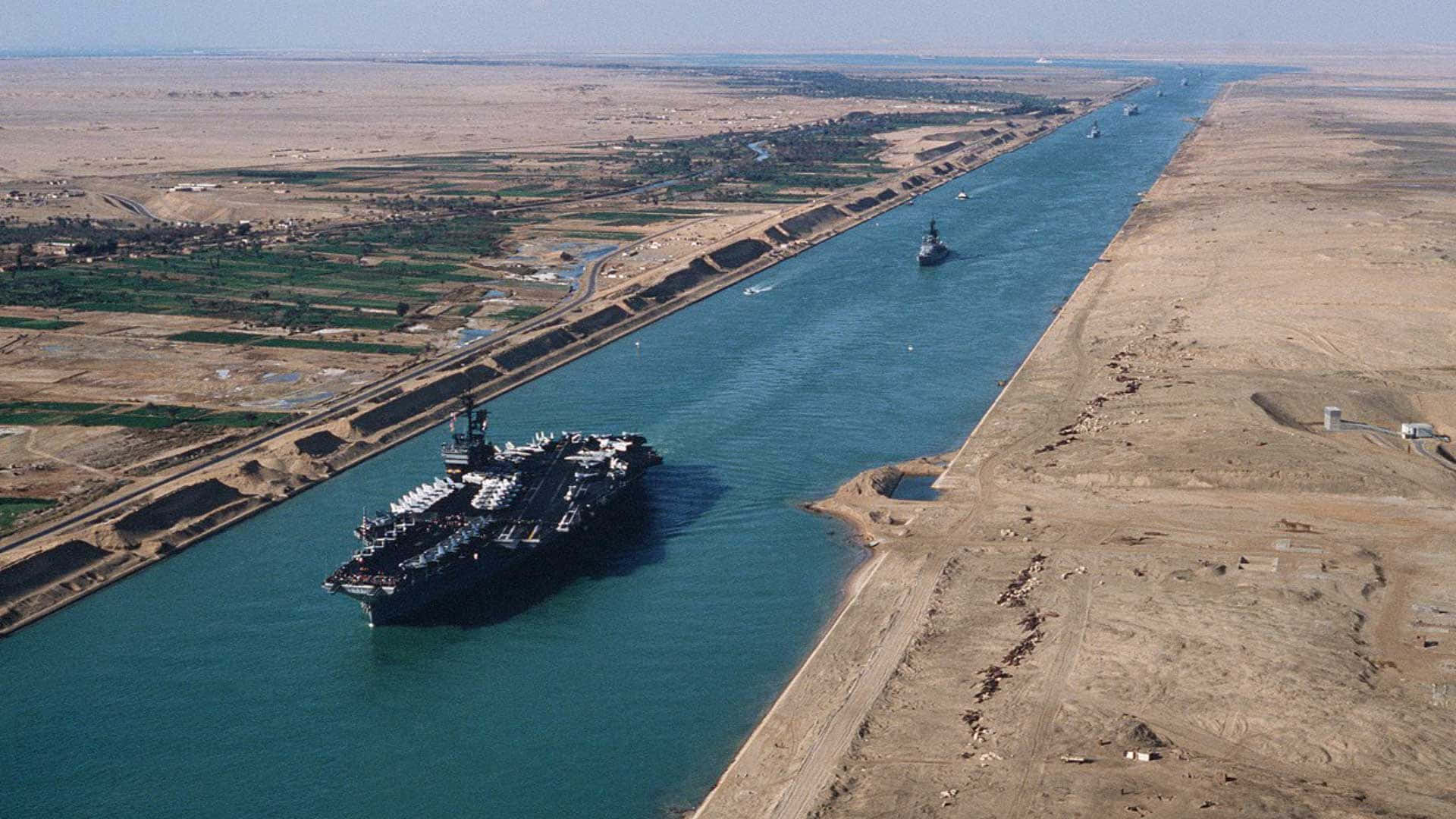 蘇伊士運河人工水道美圖 壁紙 — 一艘巨大的航空母艦在蘇伊士運河航行，兩側是乾旱的沙漠和綠色的農業地帶，天空晴朗。