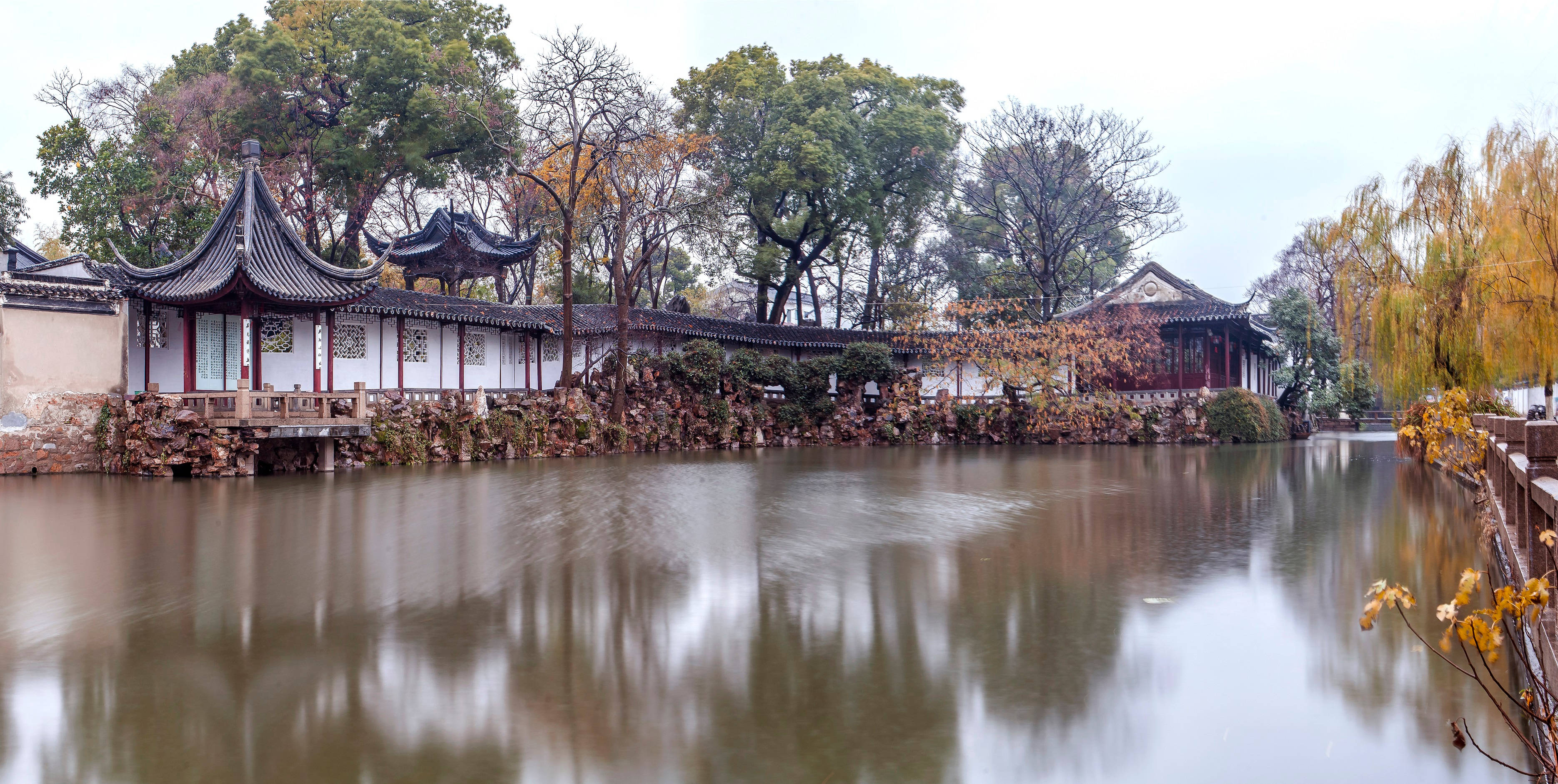 蘇州古典園林全景 壁紙 — 傳統中式亭閣的彎曲屋頂倒映在平靜的水面上，周圍環繞著秋樹。