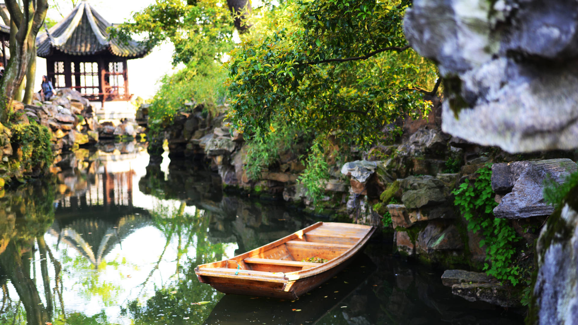 蘇州園林船景 壁紙 — 一艘木船平靜地停泊在寧靜的運河上，周圍環繞著茂密的綠植和傳統中式亭閣的倒影。