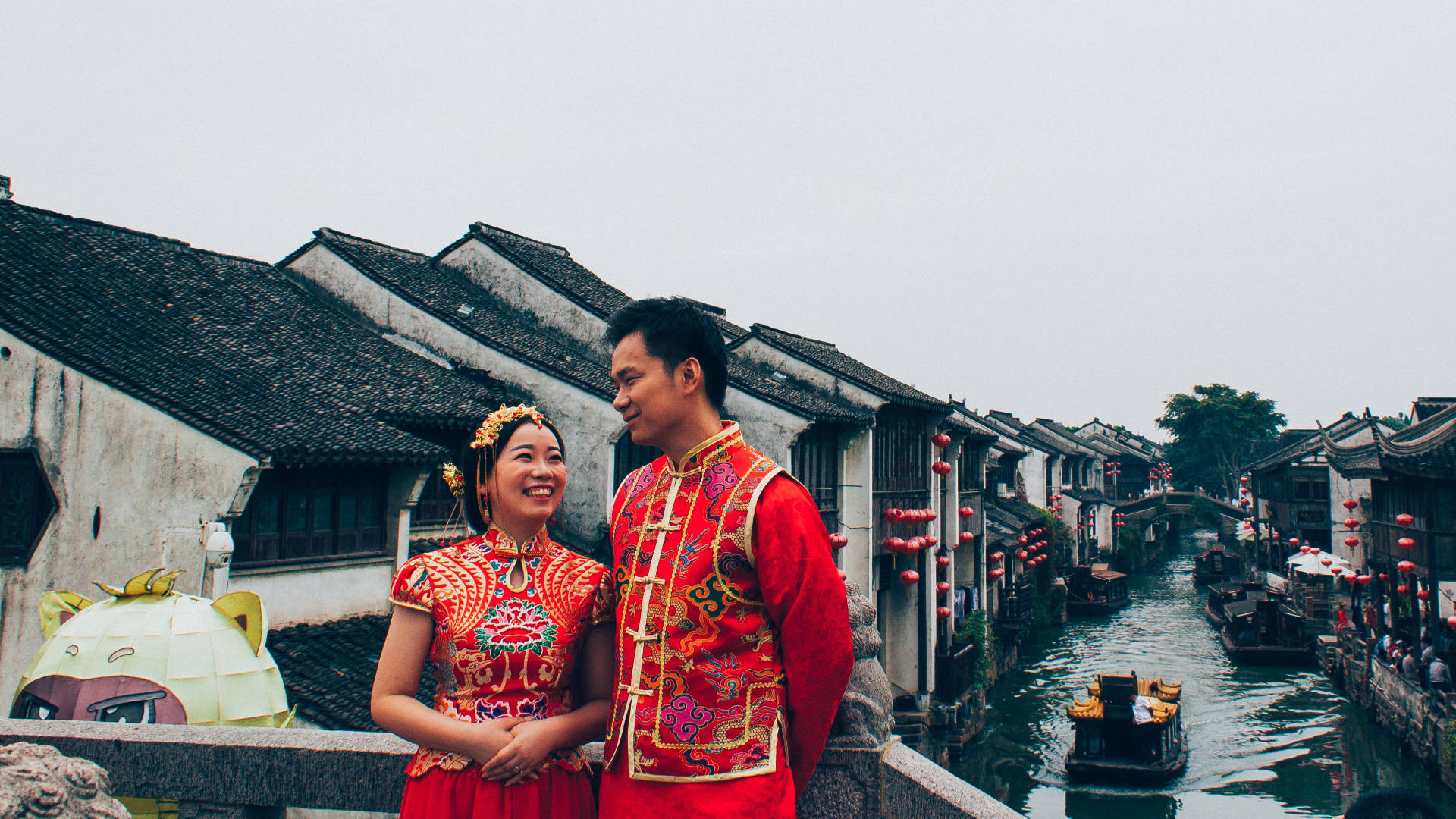 蘇州情侶合影 壁紙 — 一對微笑的新人穿著鮮豔的紅色傳統婚禮服飾，站在一座石橋上，俯瞰著兩旁歷史悠久的瓦頂房屋和漂浮船隻的運河。