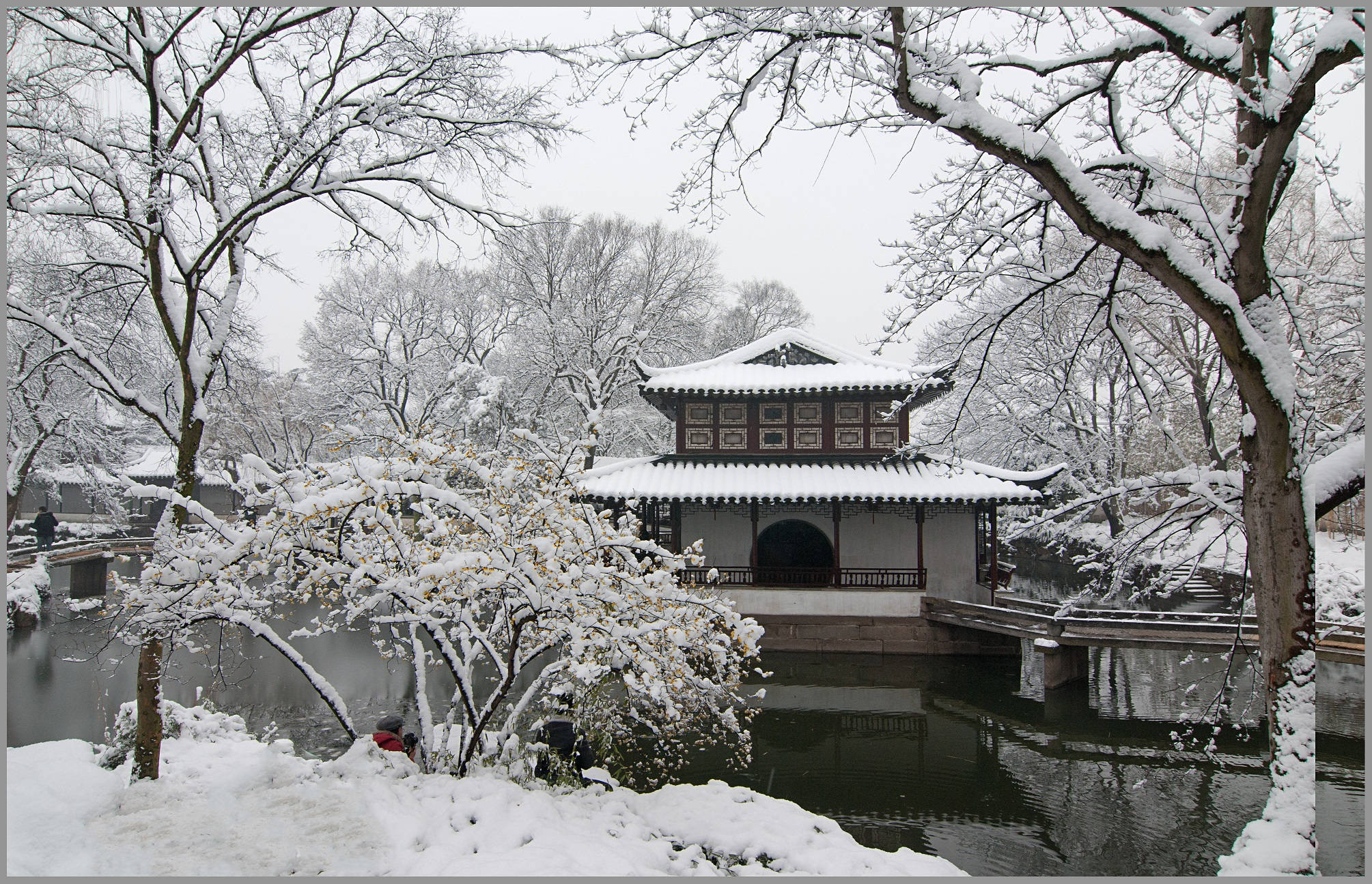 蘇州拙政園壯麗景觀 壁紙 — 一座傳統中式亭閣在雪中靜立，被雪壓枝頭的樹木環繞，旁邊是一池平靜的湖水。