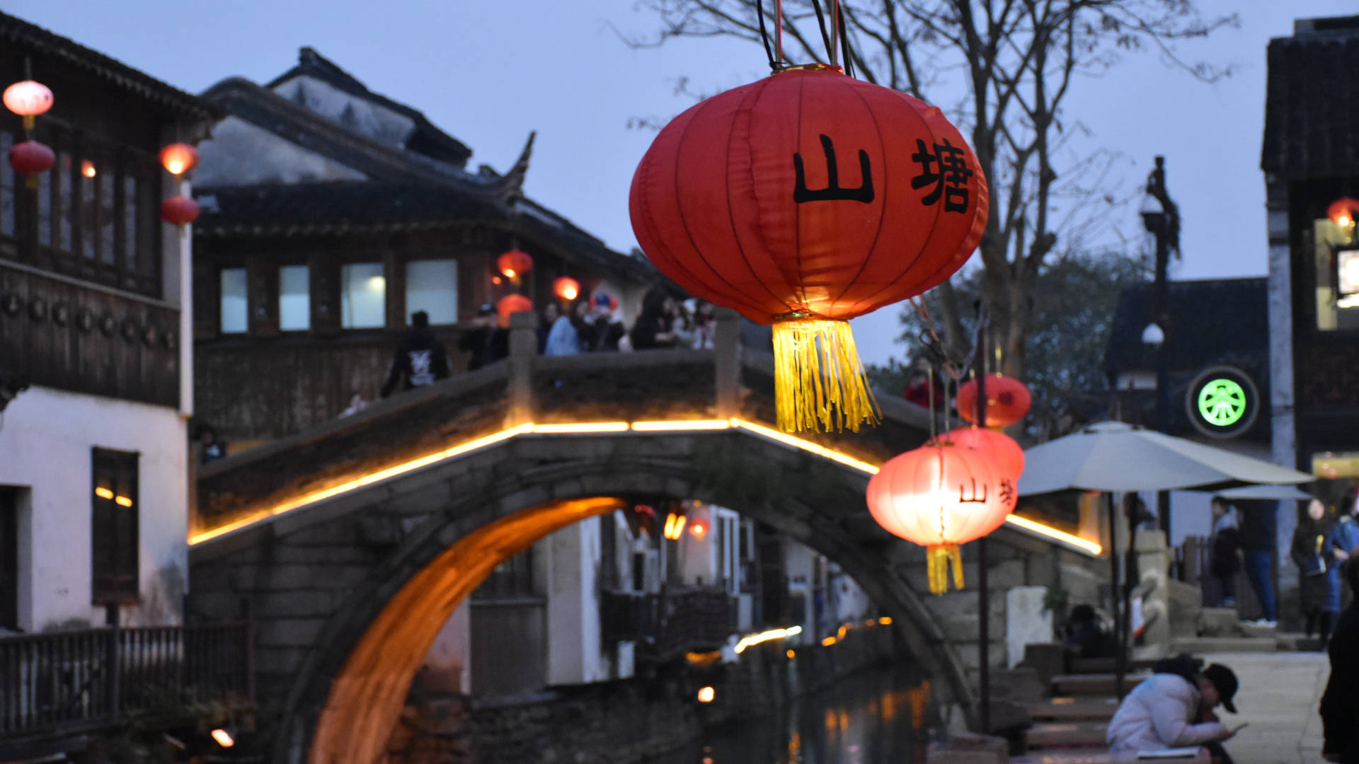 蘇州普濟橋上的紅燈籠 壁紙 — 一座石拱橋橫跨運河，在黃昏時分被掛在繁華傳統街道上方、帶有中文字樣的紅色發光燈籠照亮。
