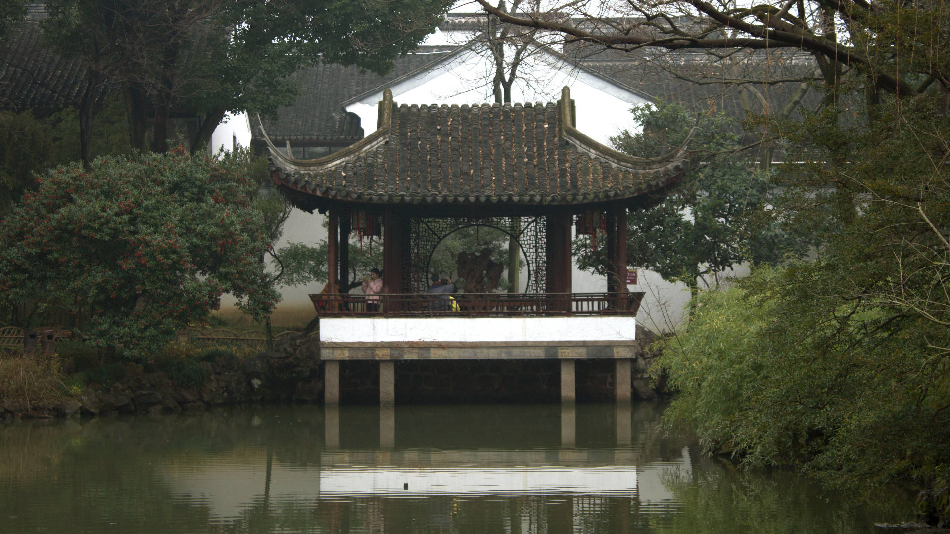 蘇州獨亭 壁紙 — 一座傳統中式亭閣，擁有彎曲屋頂，立於平靜水面的支柱上，四周環繞著茂密的綠植和光禿的樹木。