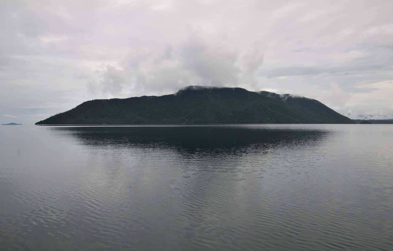 蘇拉威西島的島嶼風光 壁紙 — 一座孤獨的森林島嶼從平靜的灰色水域中升起，天空陰沉，薄霧籠罩著山頂。