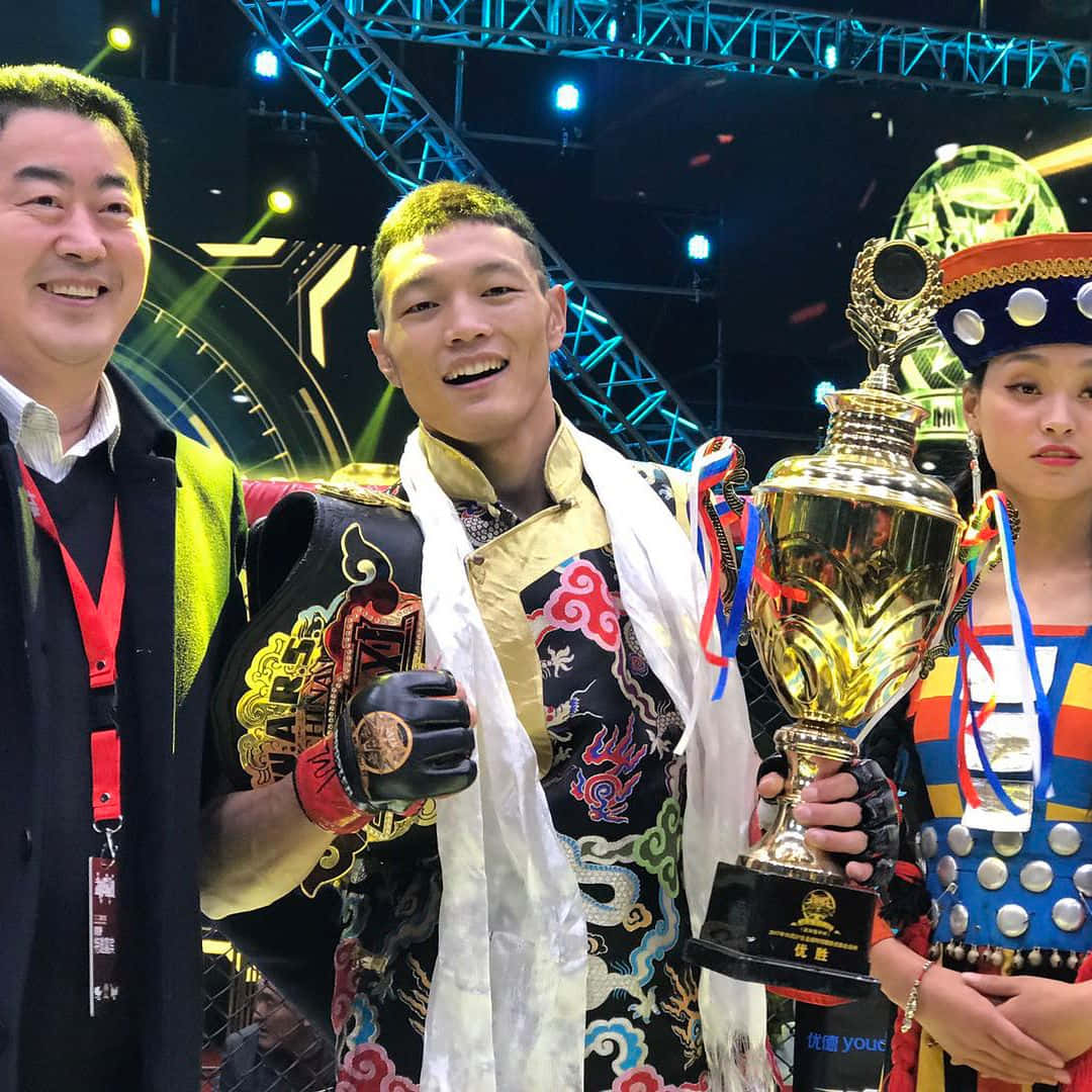 蘇木代吉手持獎盃 壁紙 — 一位身穿華麗傳統服飾的勝利戰士，在競技場燈光下，手持冠軍獎盃，身旁有兩位微笑的同伴，滿臉光彩。