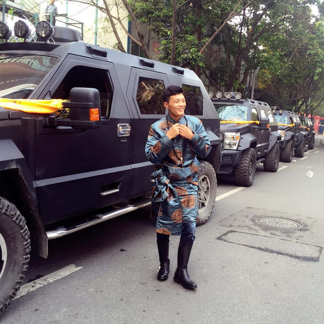 穿著藍色圖案長袍的男人在街上微笑站在黑色裝甲車旁。