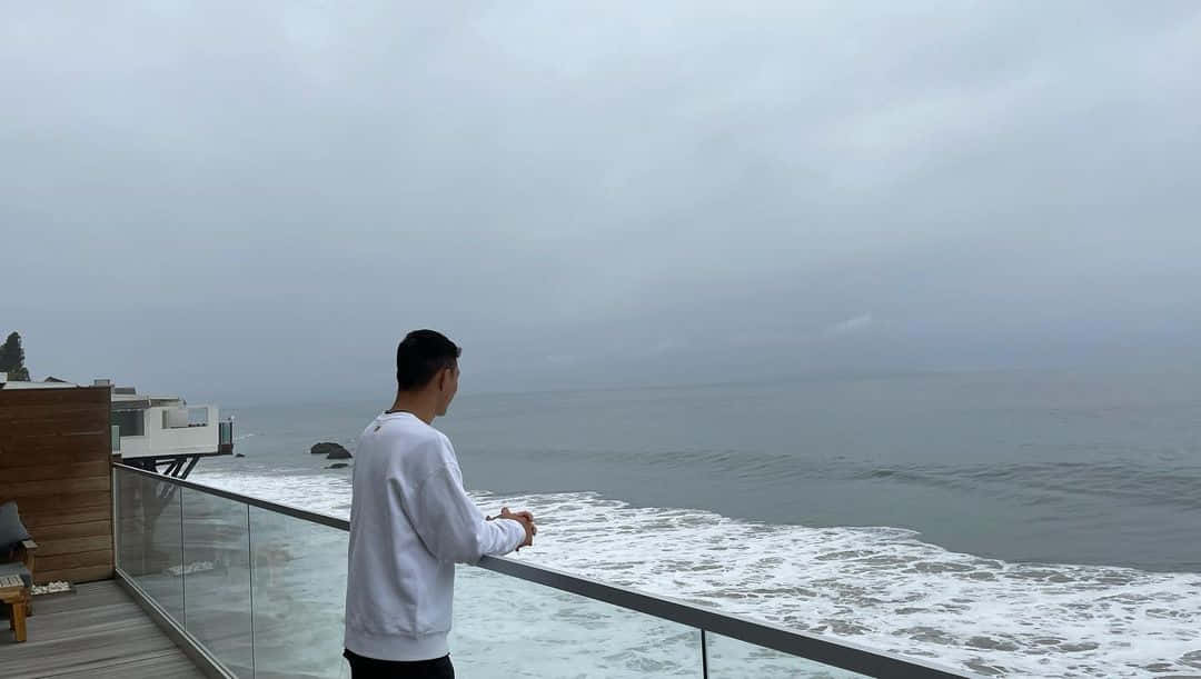 身穿白色運動衫的男子在多雲的日子裡從現代玻璃陽台凝視海洋。
