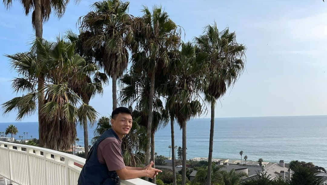 男子在陽台上微笑，身後是海洋和棕櫚樹