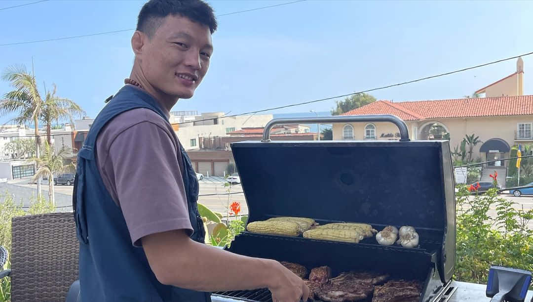 男人在陽光明媚的露臺上烤牛排和玉米，有棕櫚樹和房屋。