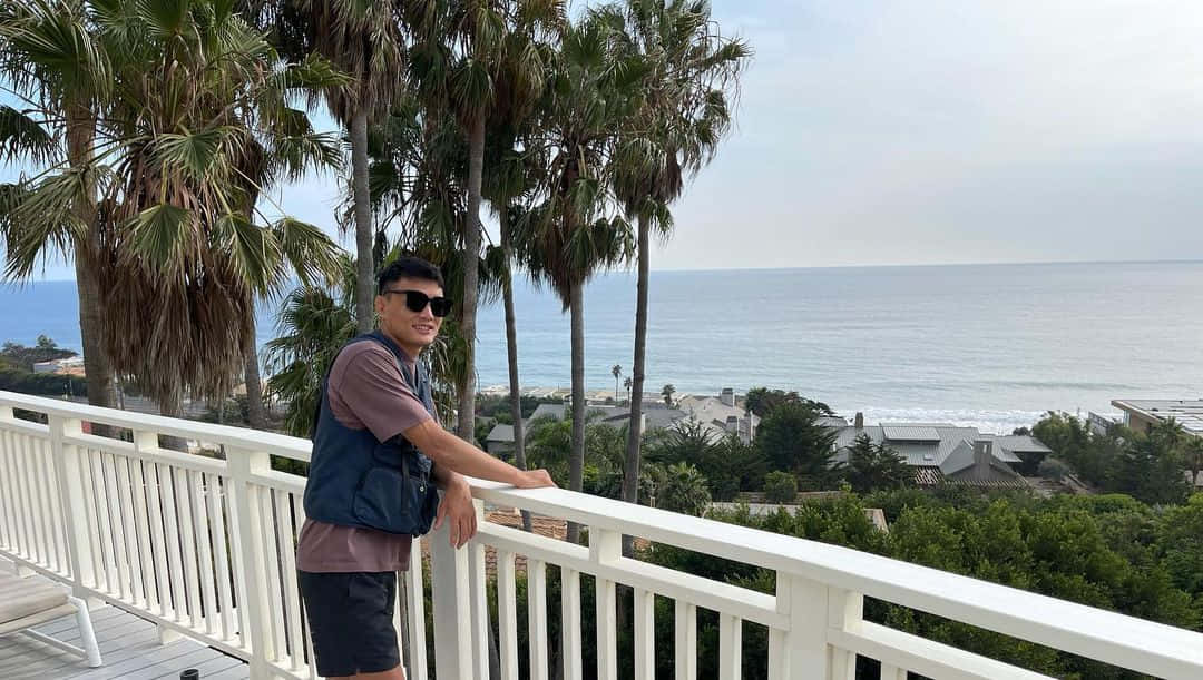 戴太陽眼鏡的男子靠在陽台欄杆上，俯瞰海洋和棕櫚樹。