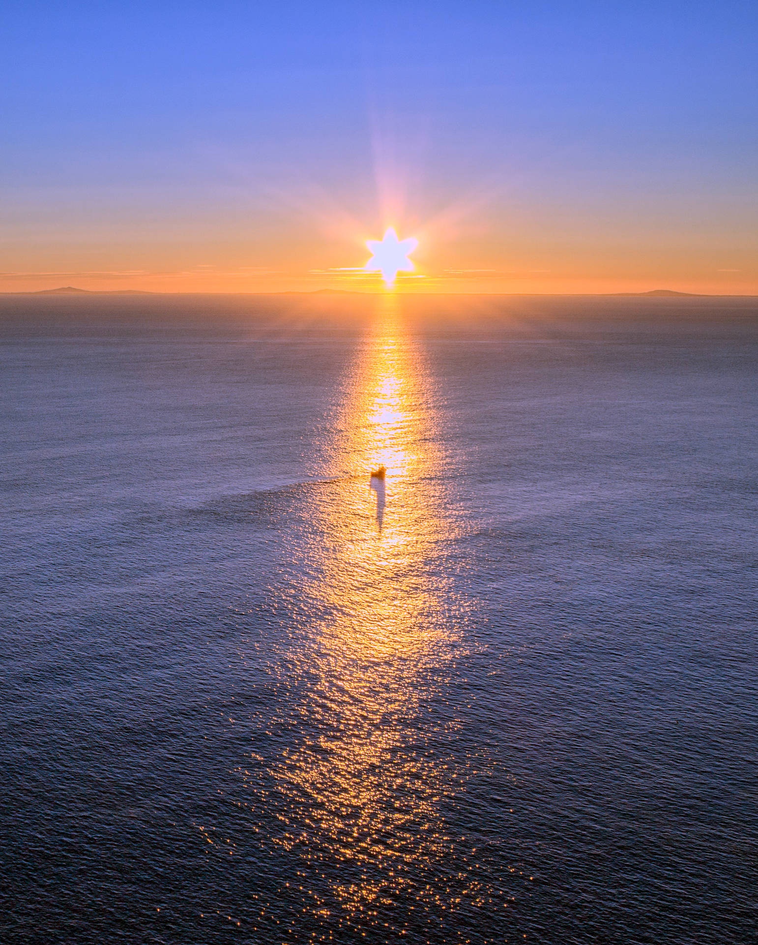 日落時平靜海面上的帆船，星形太陽投射金色倒影