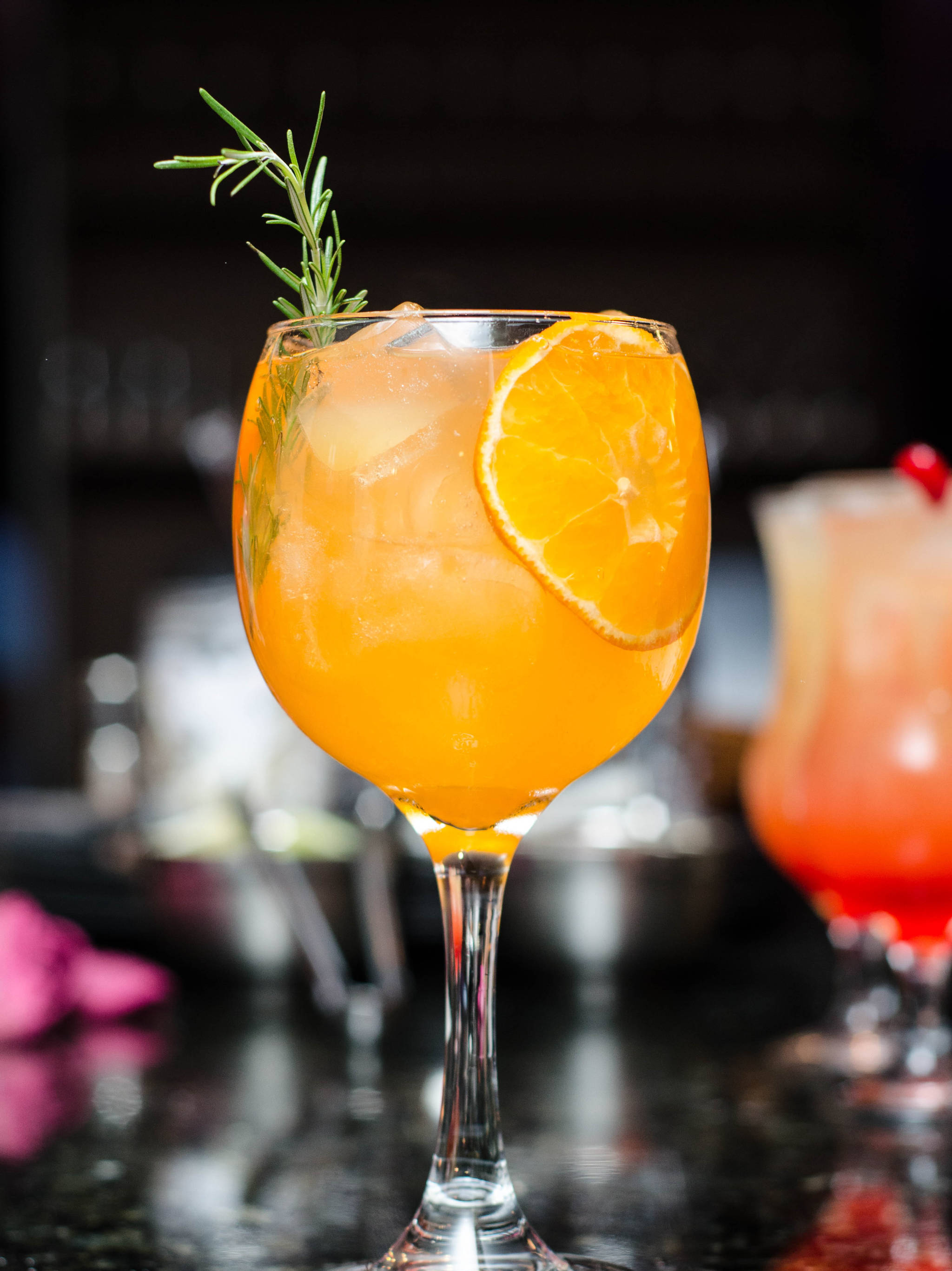 蘇格蘭威士忌熱帶飲品 壁紙 — 一杯充滿活力的橙色雞尾酒，盛於高腳杯中，飾以柑橘片和迷迭香枝，置於深色吧檯上。