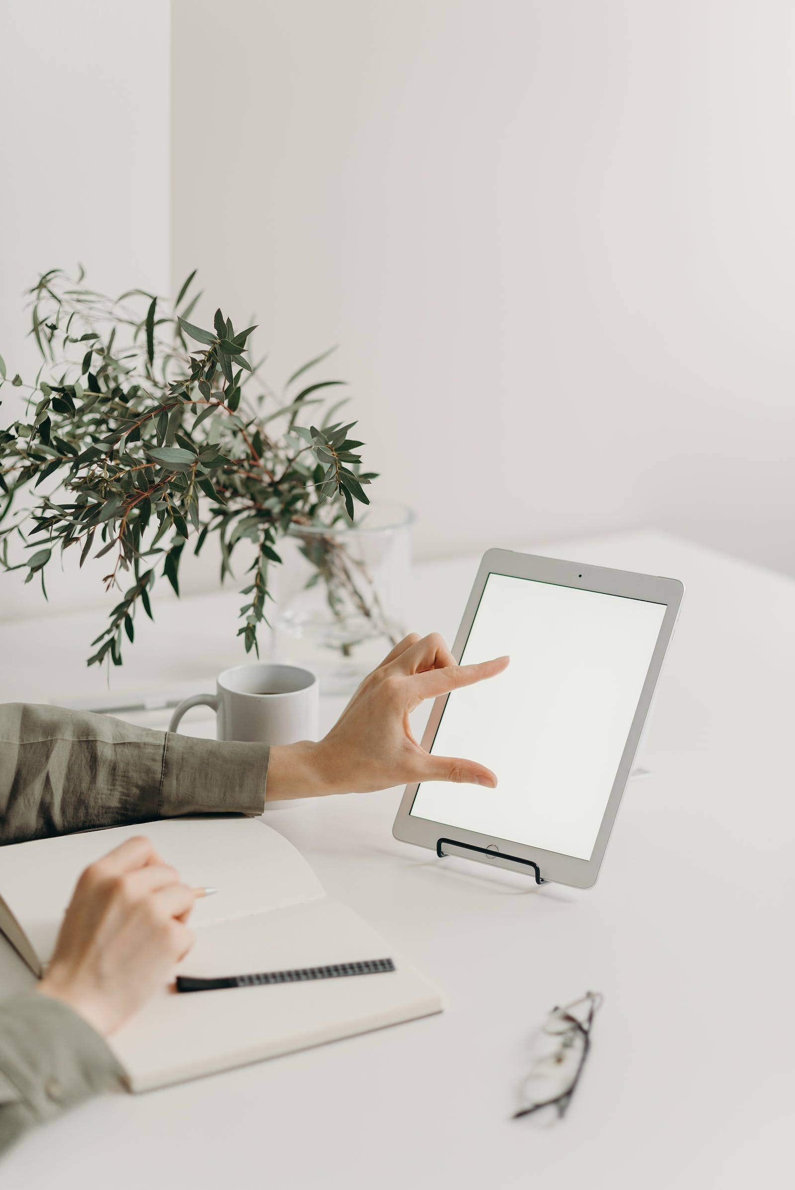 蘋果 iPad Pro 放在支架上 壁紙 — 一個人的手在簡約白色桌子上，旁邊放著綠色植物花瓶和馬克杯，一邊寫筆記本一邊操作平板電腦。