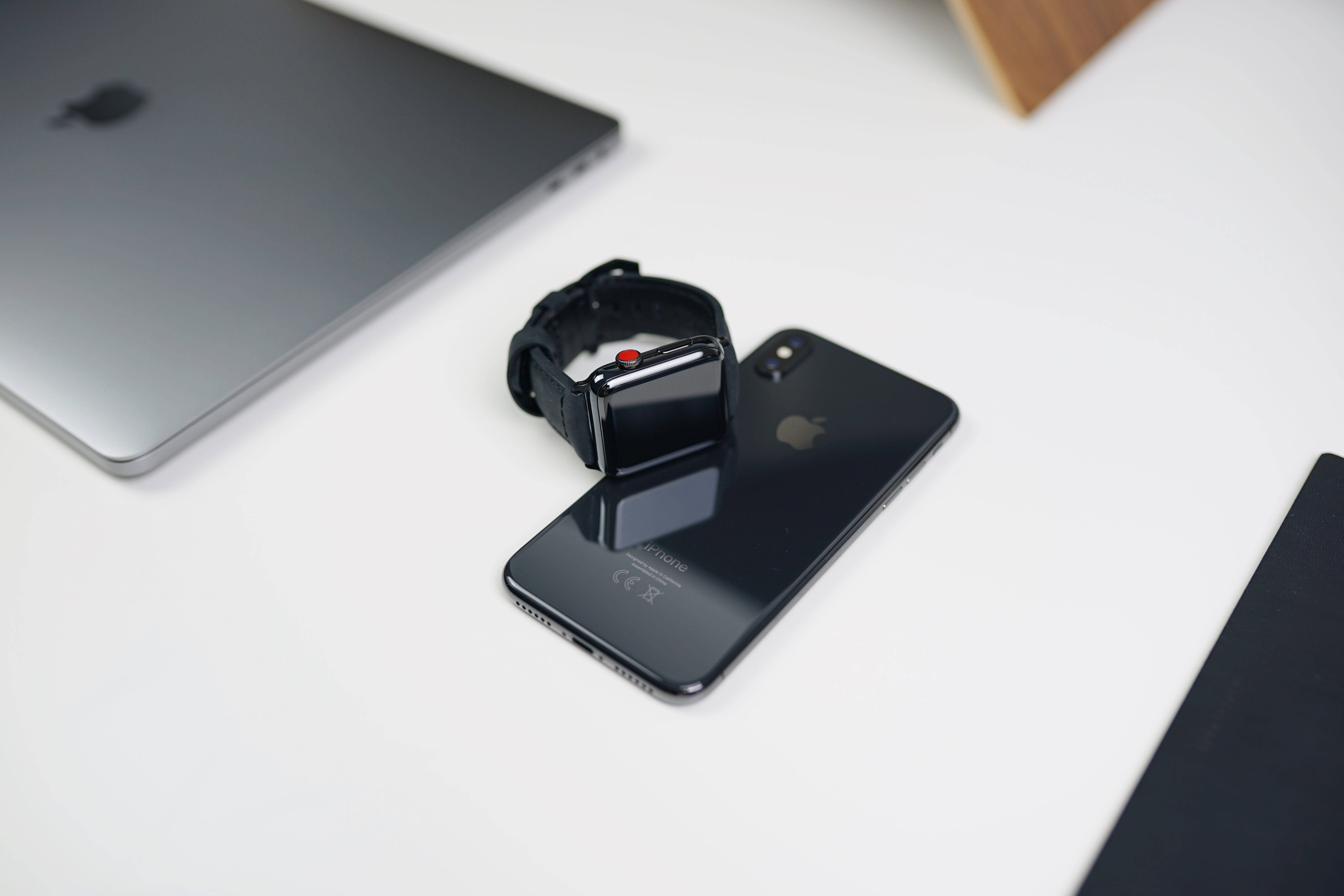 蘋果手錶與主要蘋果產品 壁紙 — 一隻時尚的黑色Apple Watch放在一部相配的iPhone上，置於簡約白色桌子上，旁邊有一部銀色手提電腦。