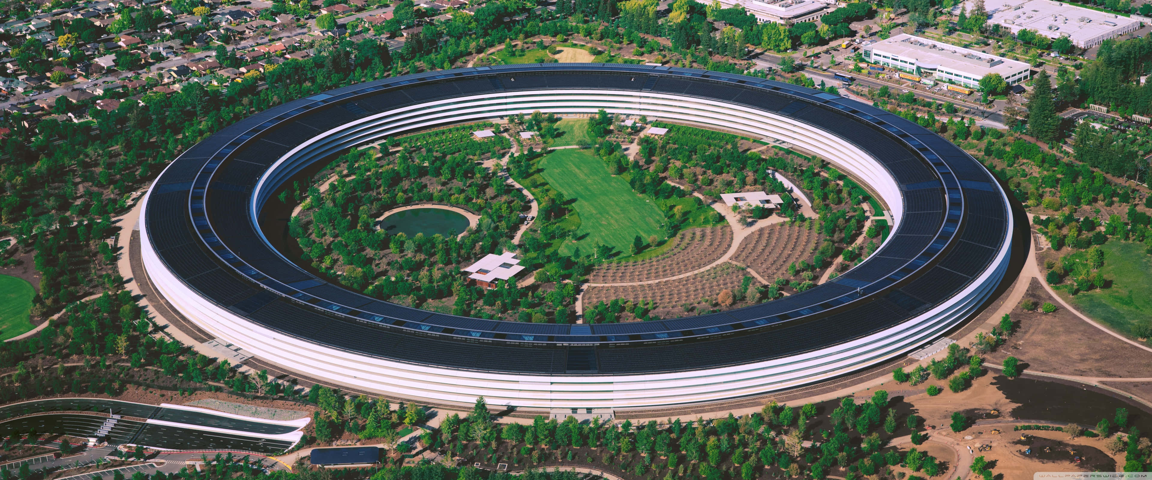 蘋果標誌在多彩星雲背景上 壁紙 — Apple Park 標誌性圓形太空船建築的鳥瞰圖，周圍環繞著茂密的綠化和修剪整齊的草坪。