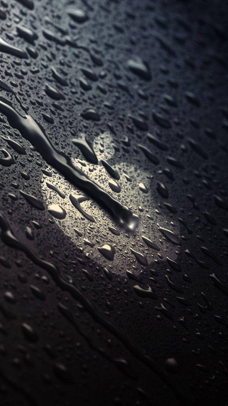 蘋果標誌水滴 iPhone SE 壁紙 — 閃爍的雨滴在聚焦光束照射的黑暗表面上傾瀉而下，營造出陰鬱的反射場景。