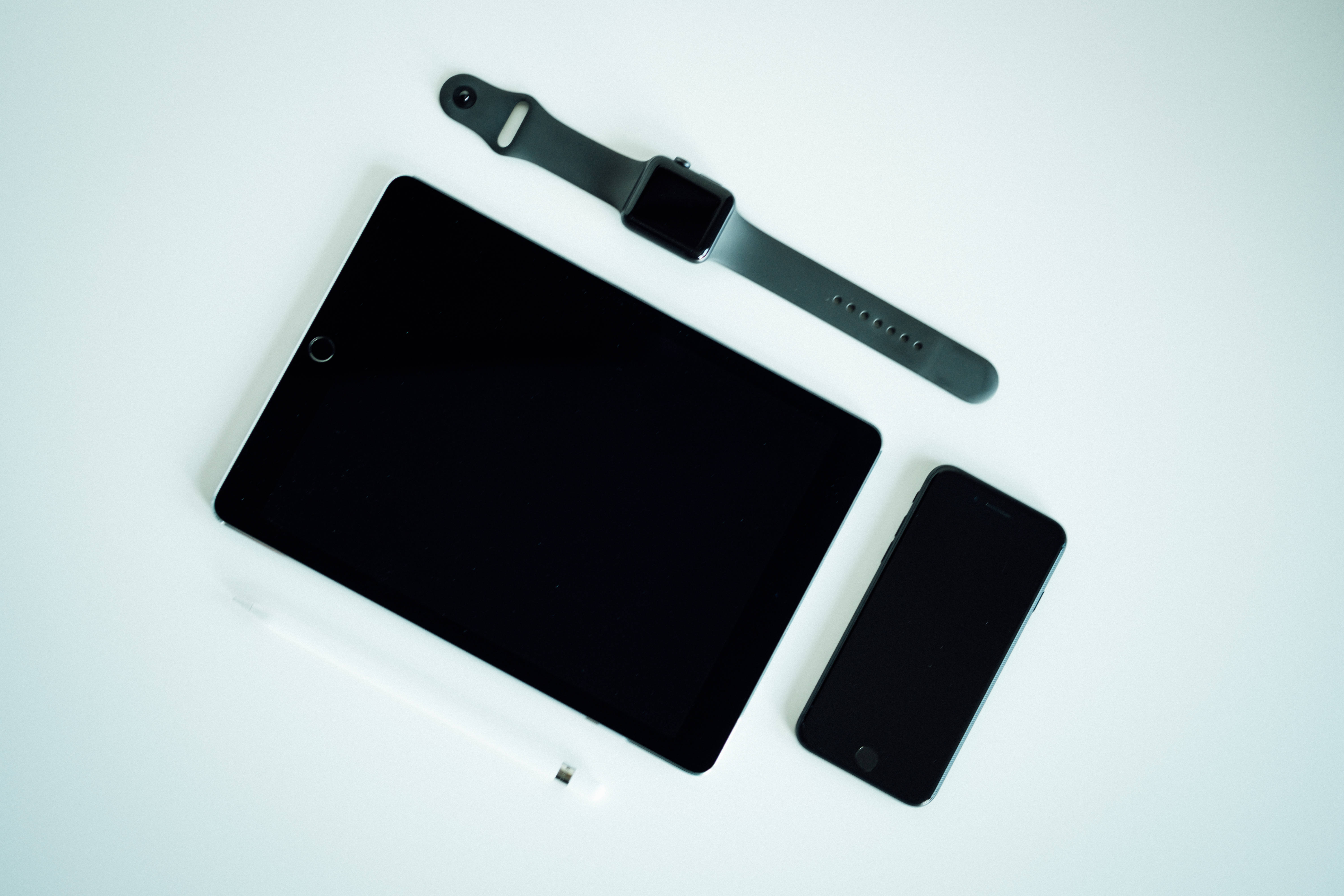 蘋果裝置與iPad Pro 壁紙 — 簡約平鋪擺拍：黑色平板電腦、智能手錶和手機，搭配白色觸控筆，置於潔白表面上。