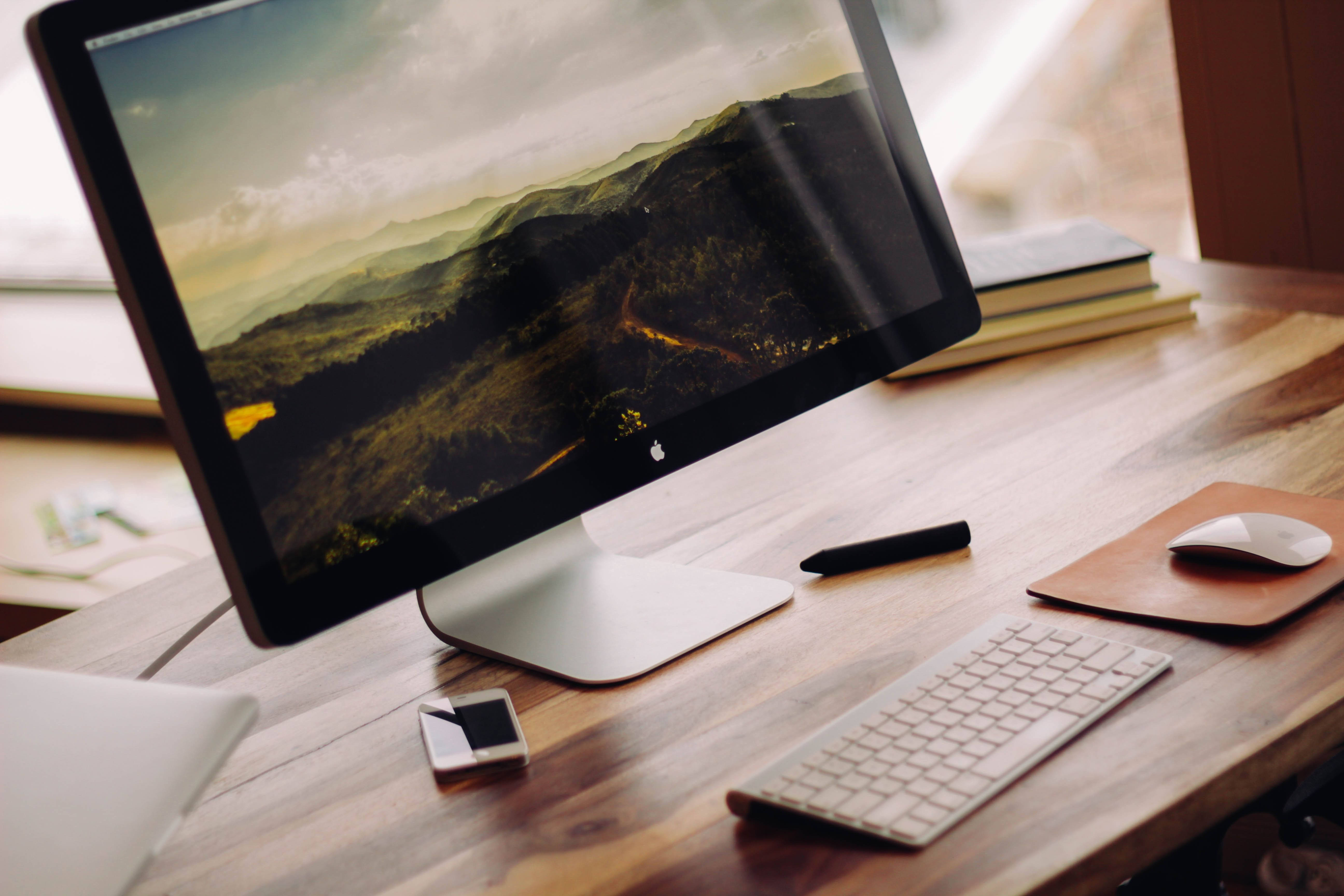 蘋果電腦擺放在木質辦公桌 壁紙 — 一部時尚的 iMac 放在木桌上，顯示著山景桌布，周圍有鍵盤、滑鼠和電話。