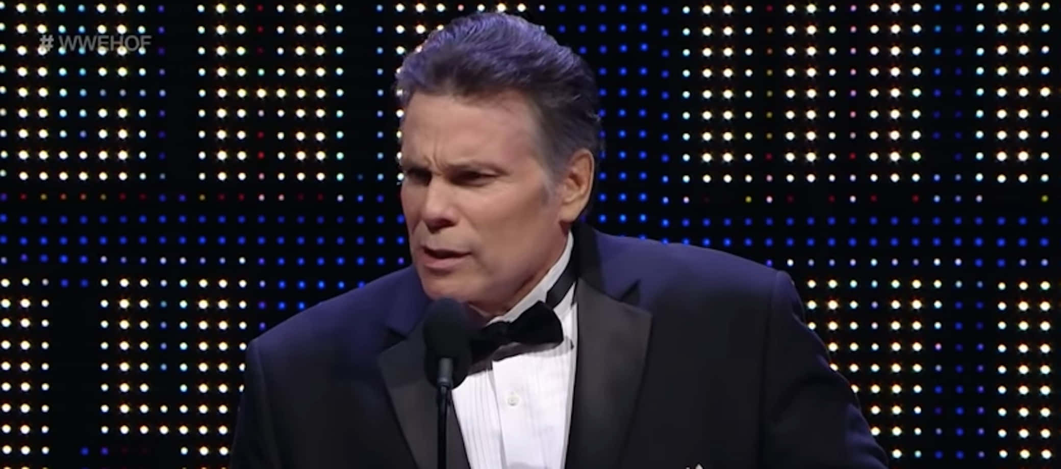 蘭尼·波福黑屏照 壁紙 — 一名身穿燕尾服的男士在發光LED點的背景前對著麥克風講話。