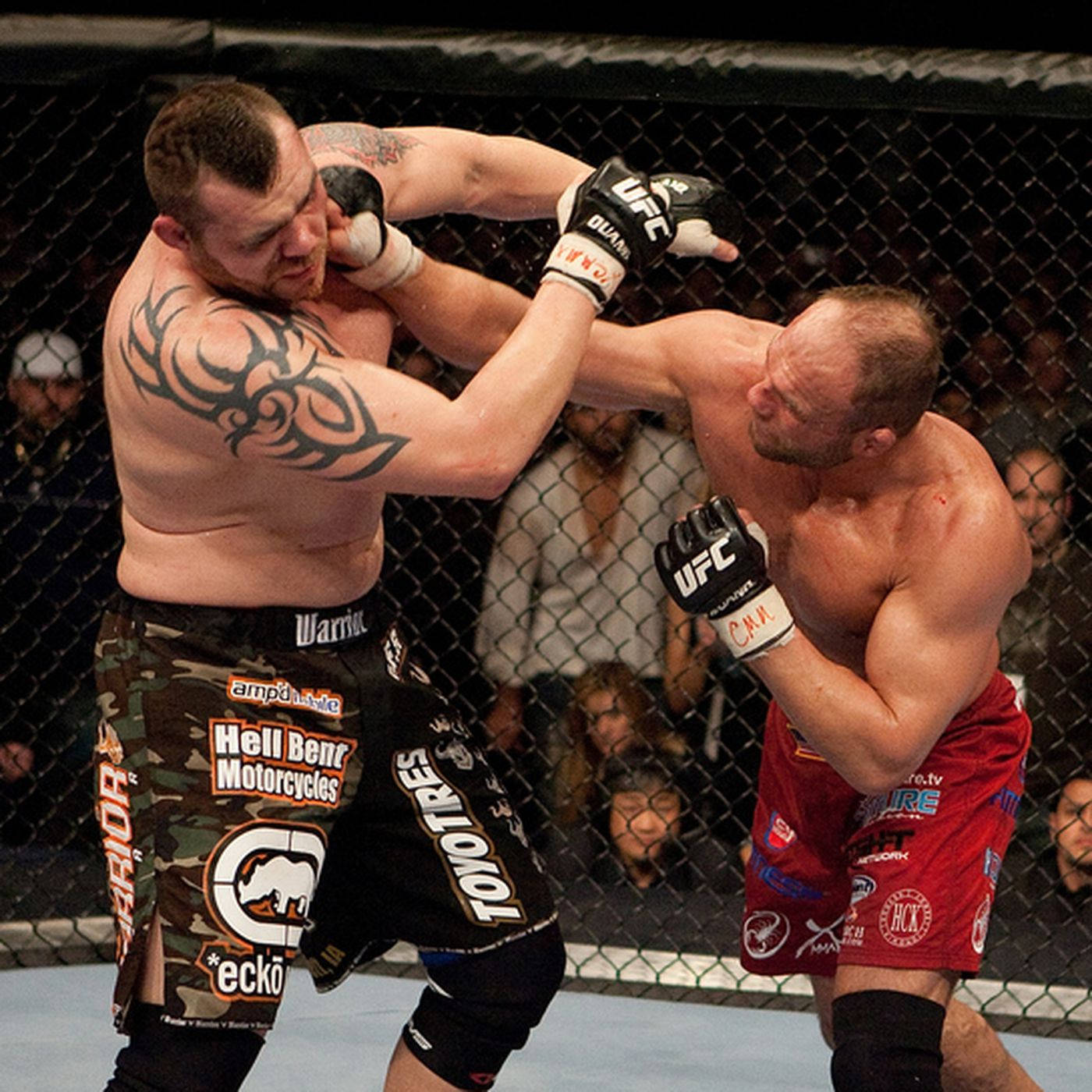 蘭迪·庫圖爾出拳 壁紙 — 兩名肌肉發達的MMA拳手在明亮的場館燈光下於八角籠內激烈纏鬥。