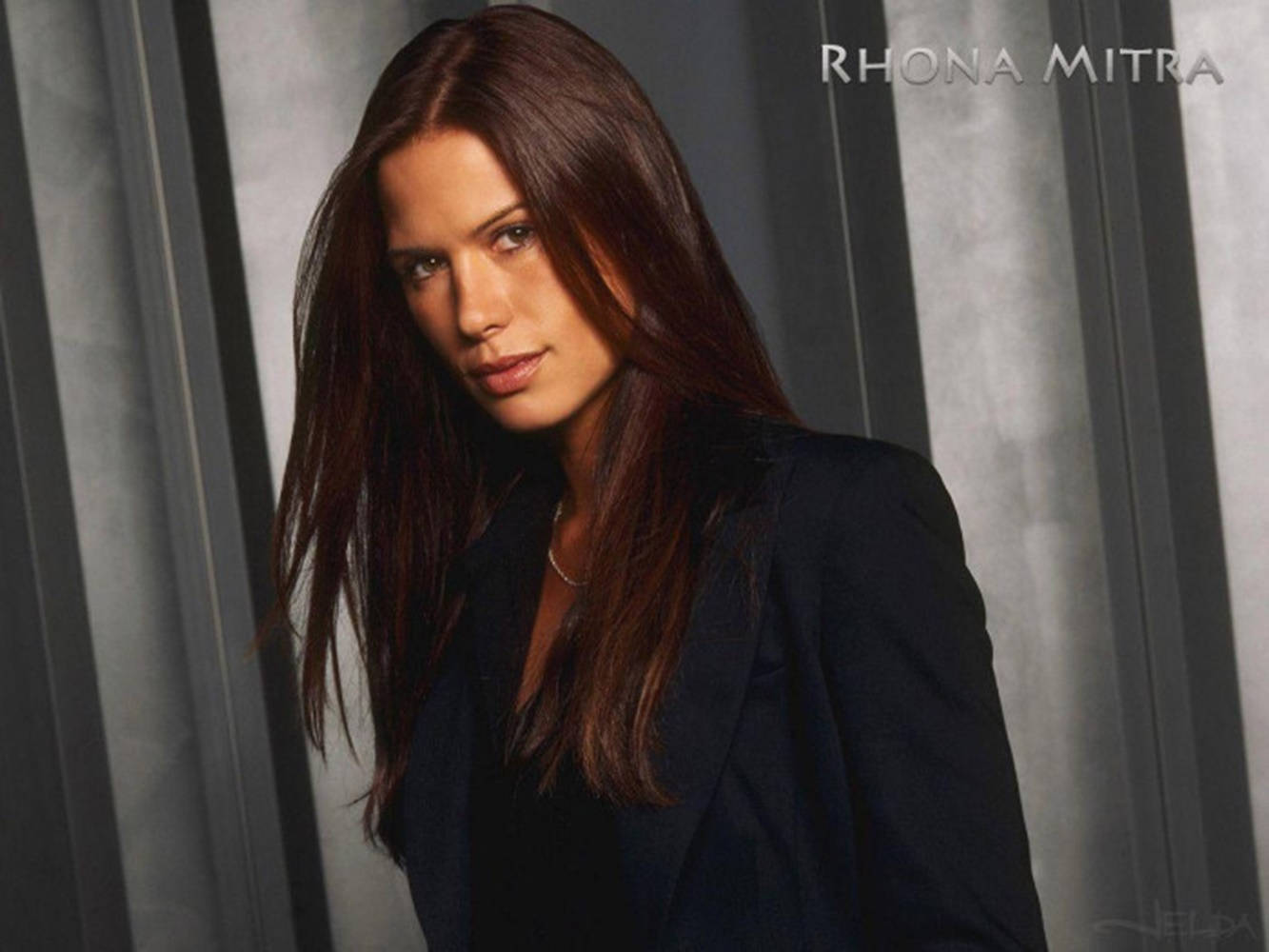 Rhona Mitra 身穿深色西裝外套，背景為灰色垂直條紋。