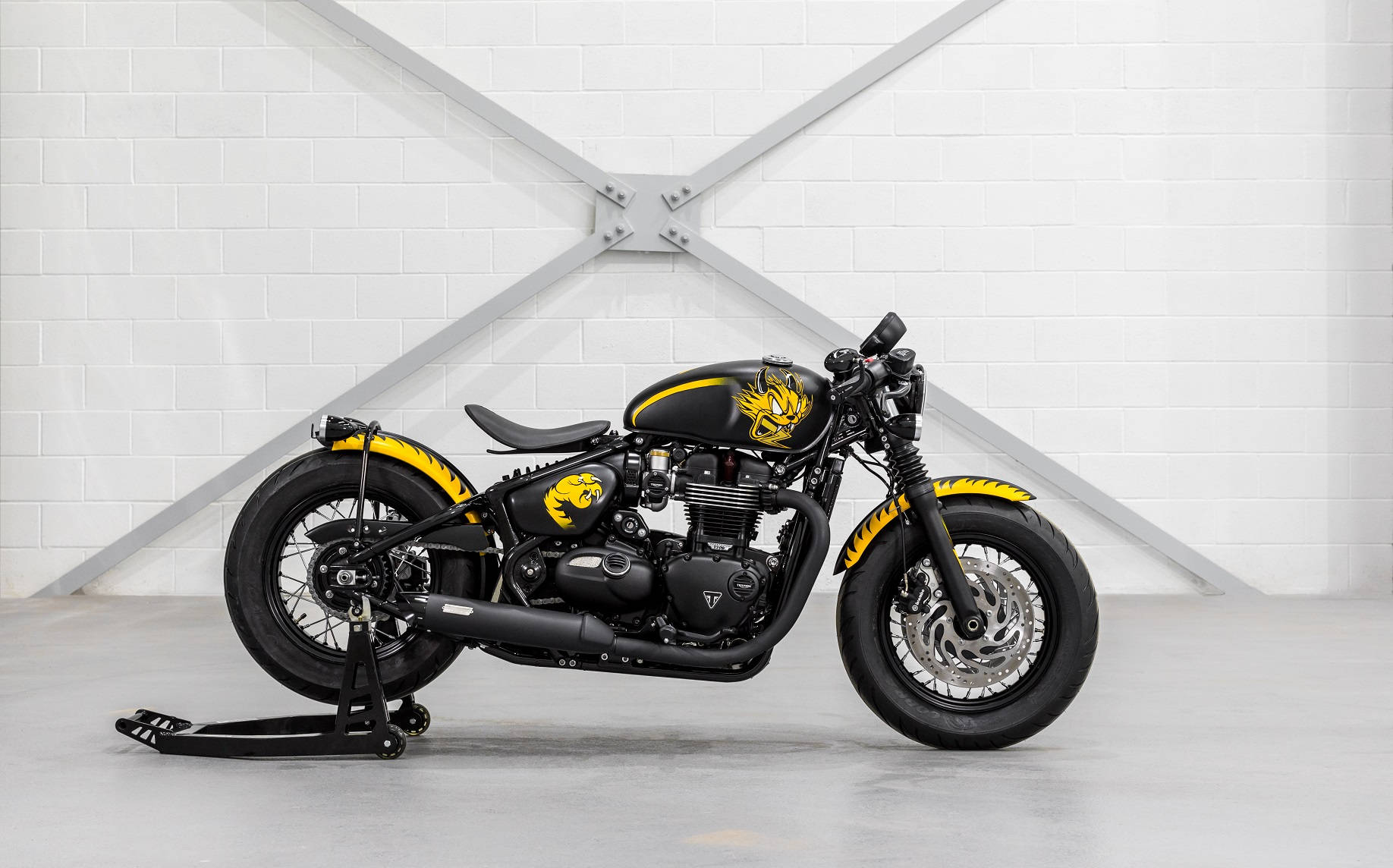 虎紋黑 Bobber Triumph Bonneville 定製版 壁紙 — 一架時尚黑色電單車帶有搶眼的黃色火焰貼紙，停放在支架上，背景是帶有金屬橫樑的白色磚牆。