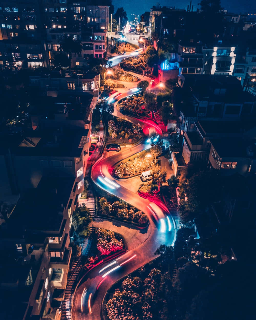 Retina Display 桌布 壁紙 — 蜿蜒城市道路的夜間空中景觀，汽車發光的燈光軌跡穿過明亮的住宅建築。