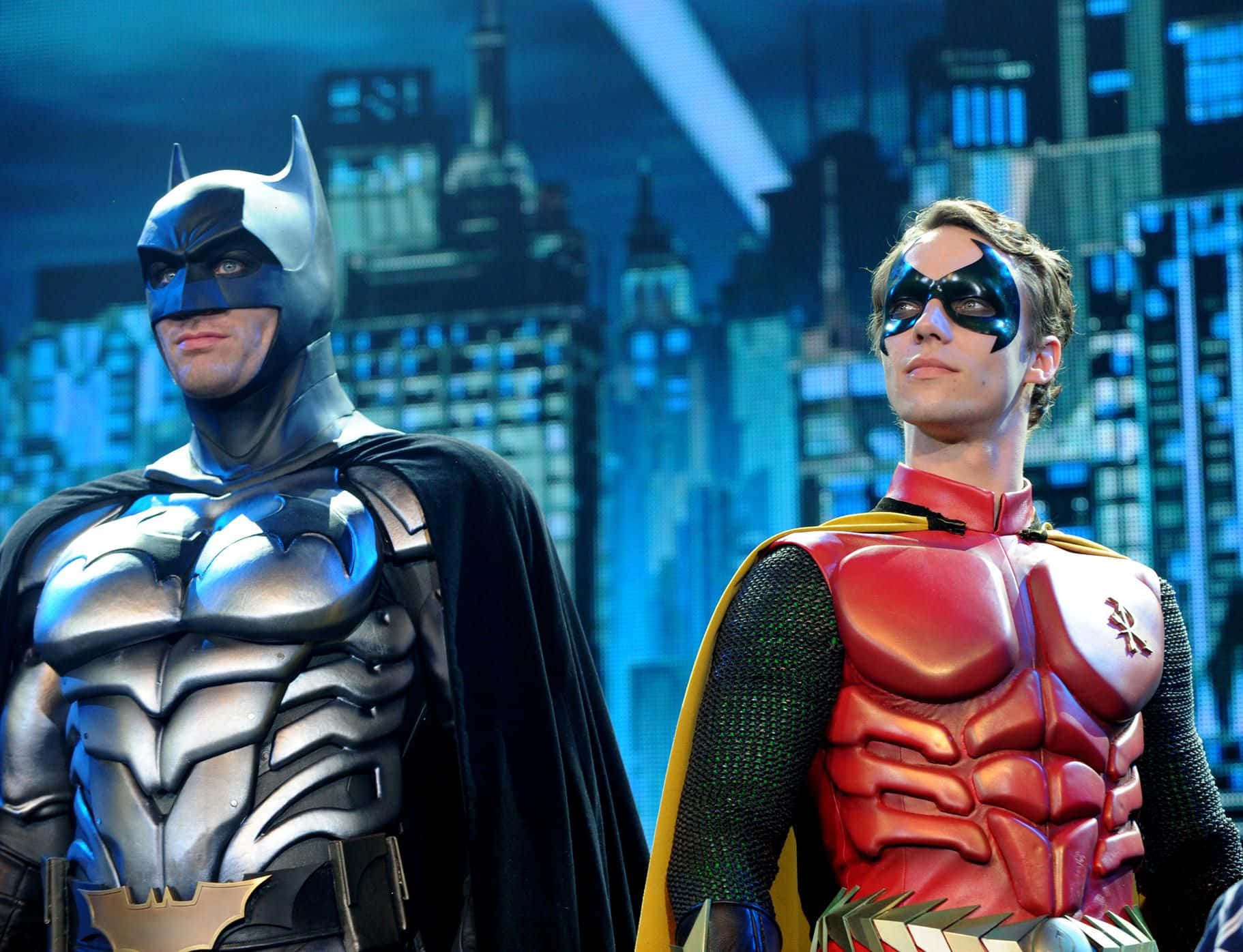 Batman Vs Robin 桌布 壁紙 — 蝙蝠俠和羅賓穿著精細的服裝並肩站立，背景是發光的藍色城市天際線。