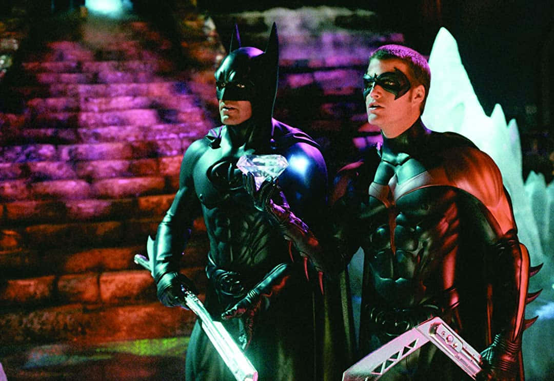 Batman Vs Robin 桌布 壁紙 — 蝙蝠俠和羅賓身穿深色服裝並肩站立，手持發光武器，背景是磚牆，被紫色和紅色的霓虹燈照亮。