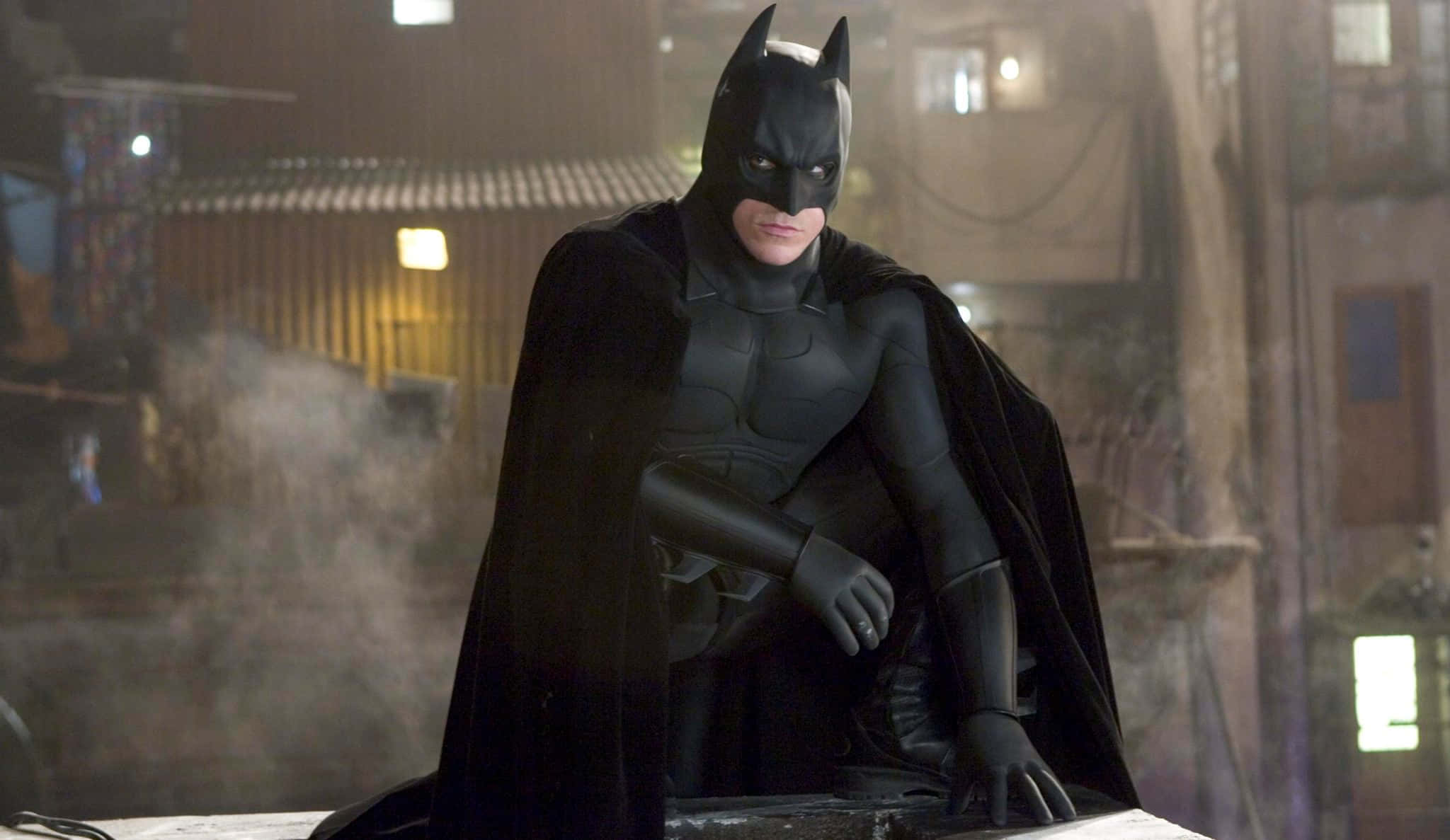 Batman Begins 桌布 壁紙 — 蝙蝠俠在夜晚蹲在黑暗煙霧瀰漫的小巷中，背景有閃爍的燈光。