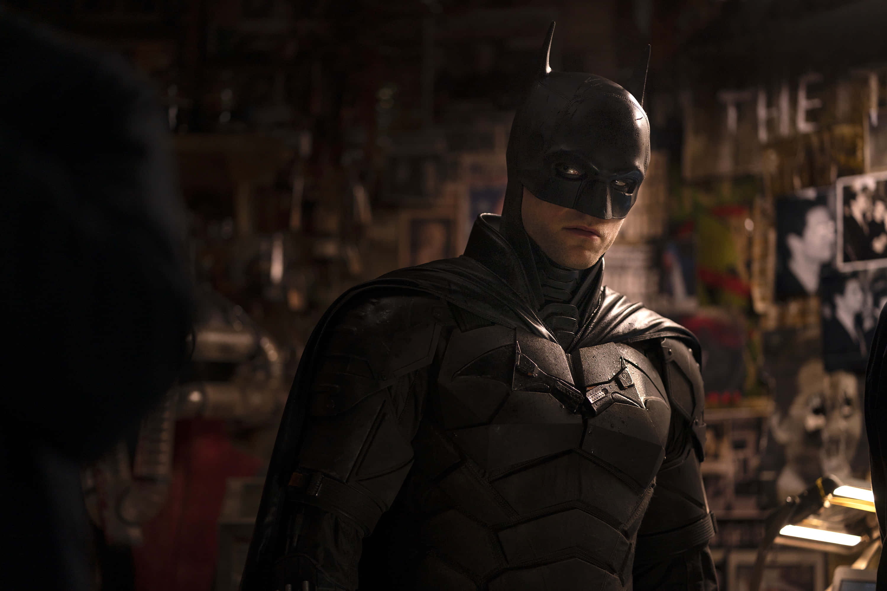 Bat-suit 桌布 壁紙 — 蝙蝠俠站在一個光線昏暗、雜亂無章的房間裡，房間裡充滿了海報和紀念品，他的裝甲服在聚焦的光線下閃閃發光。