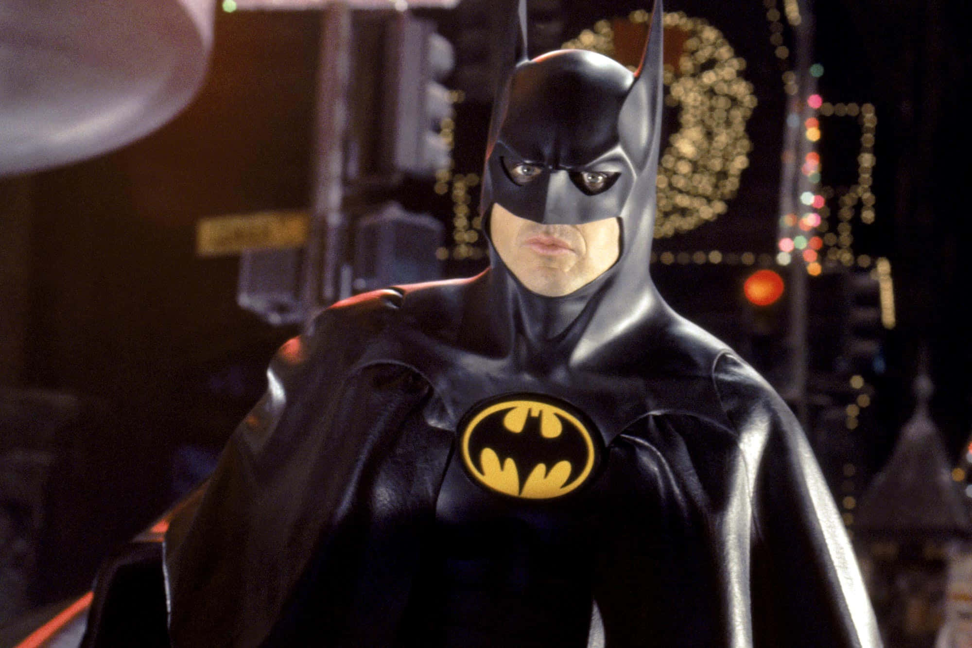 Bat-suit 桌布 壁紙 — 蝙蝠俠站在黑暗的城市景觀中，身後有發光的散景燈光，穿著他標誌性的黑色西裝和黃色蝙蝠標誌。