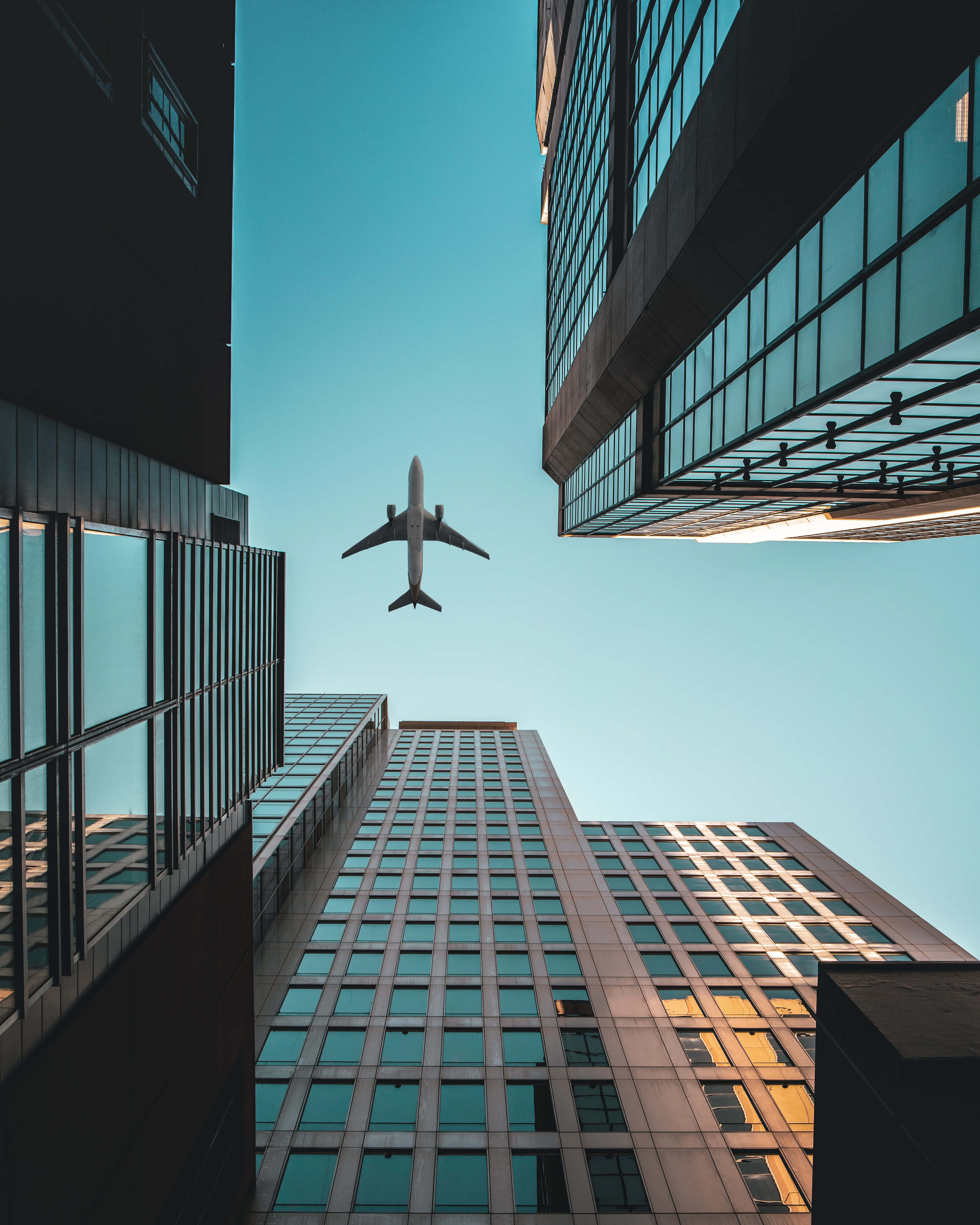 蟲眼視角建築背景 Android 飛機背景 壁紙 — 一架商用飛機在晴朗的藍天下，於高聳的玻璃摩天大樓之間翱翔。