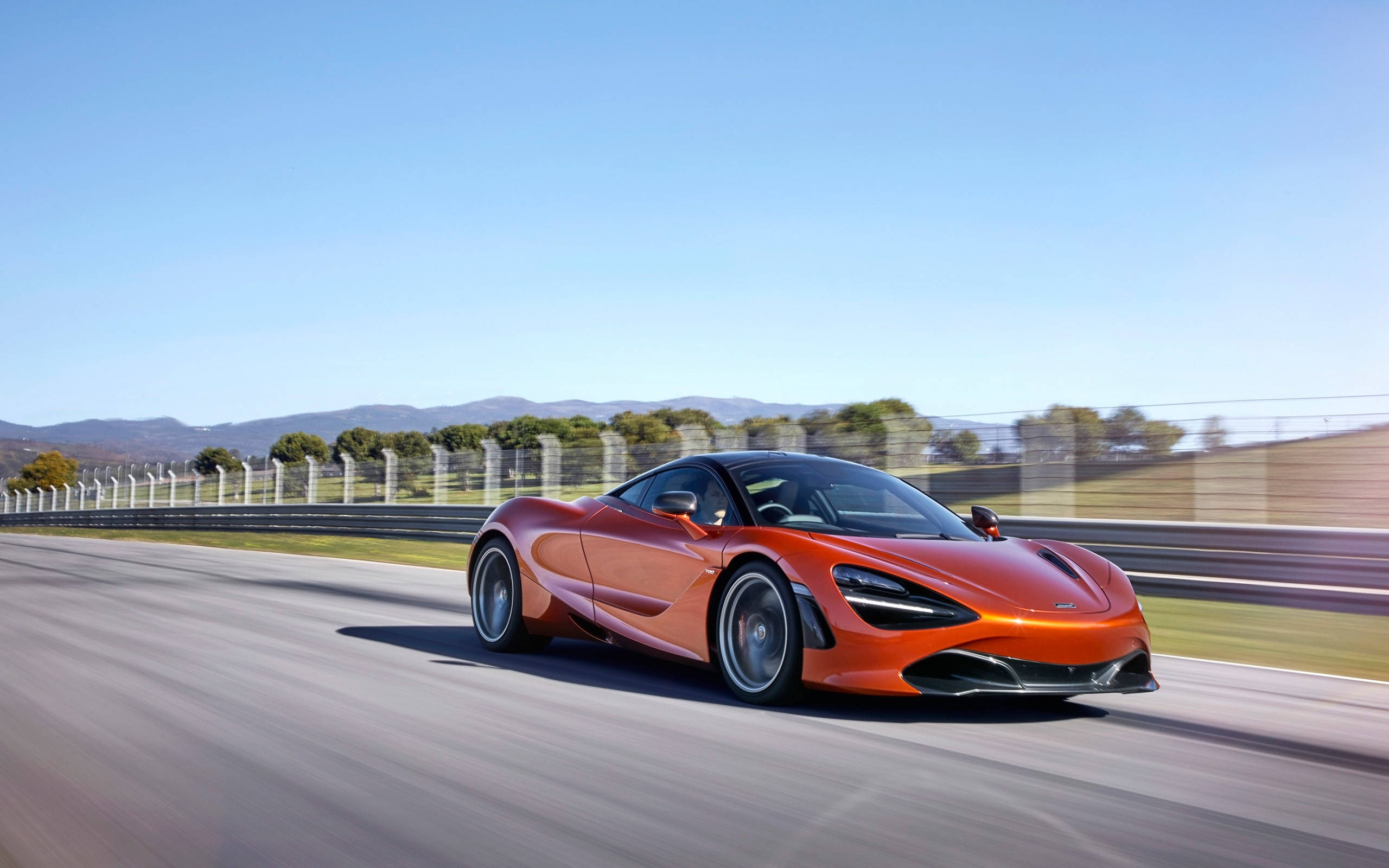 行駛中的橙色麥拉倫720s 4K 壁紙 — 一輛充滿活力的橙色McLaren跑車在晴朗的藍天下，沿著賽道高速行駛，呈現動態模糊效果。