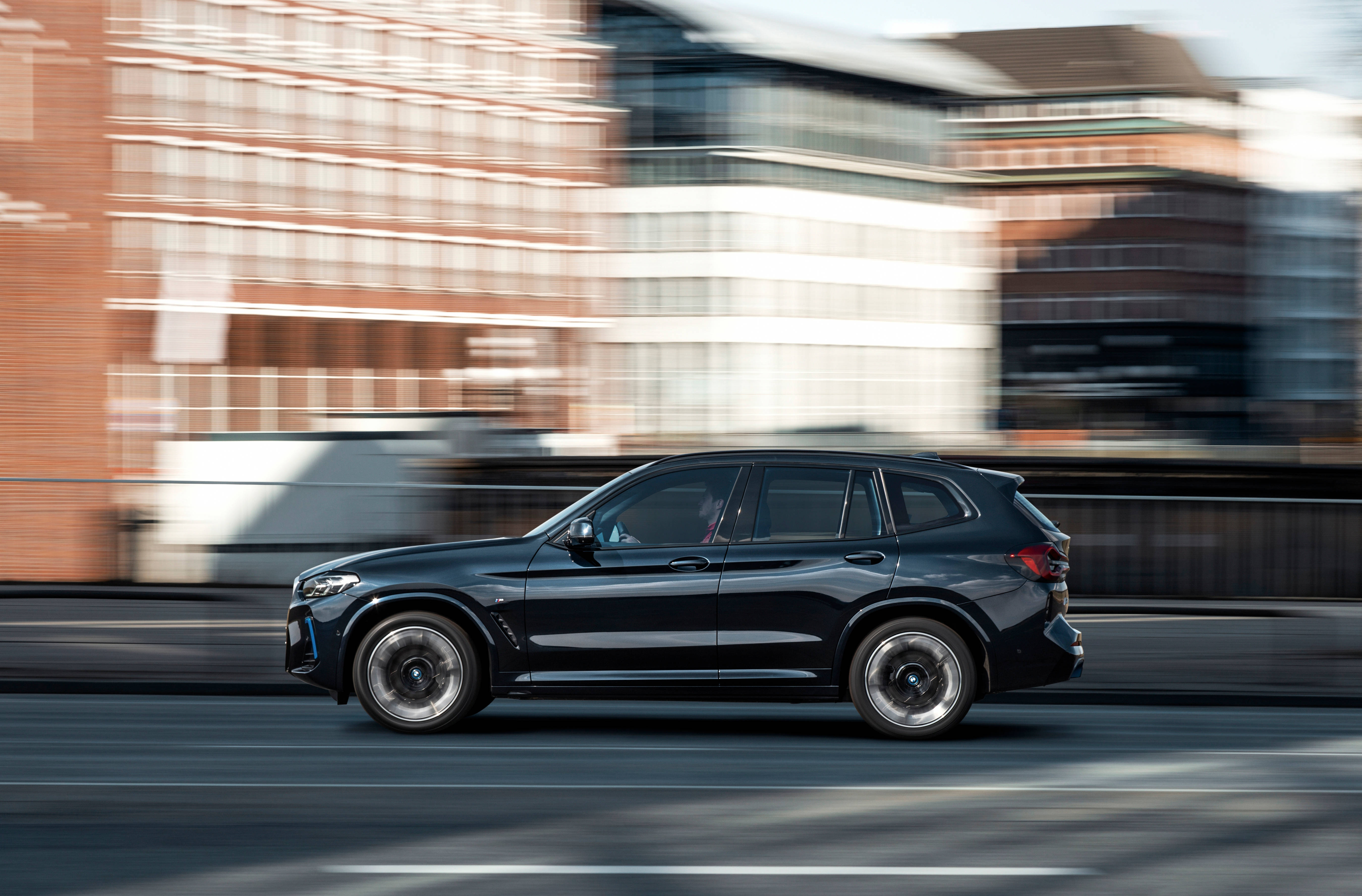 行駛中的黑色BMW 壁紙 — 一輛時尚黑色SUV在明亮的日光下高速駛過模糊的城市建築。