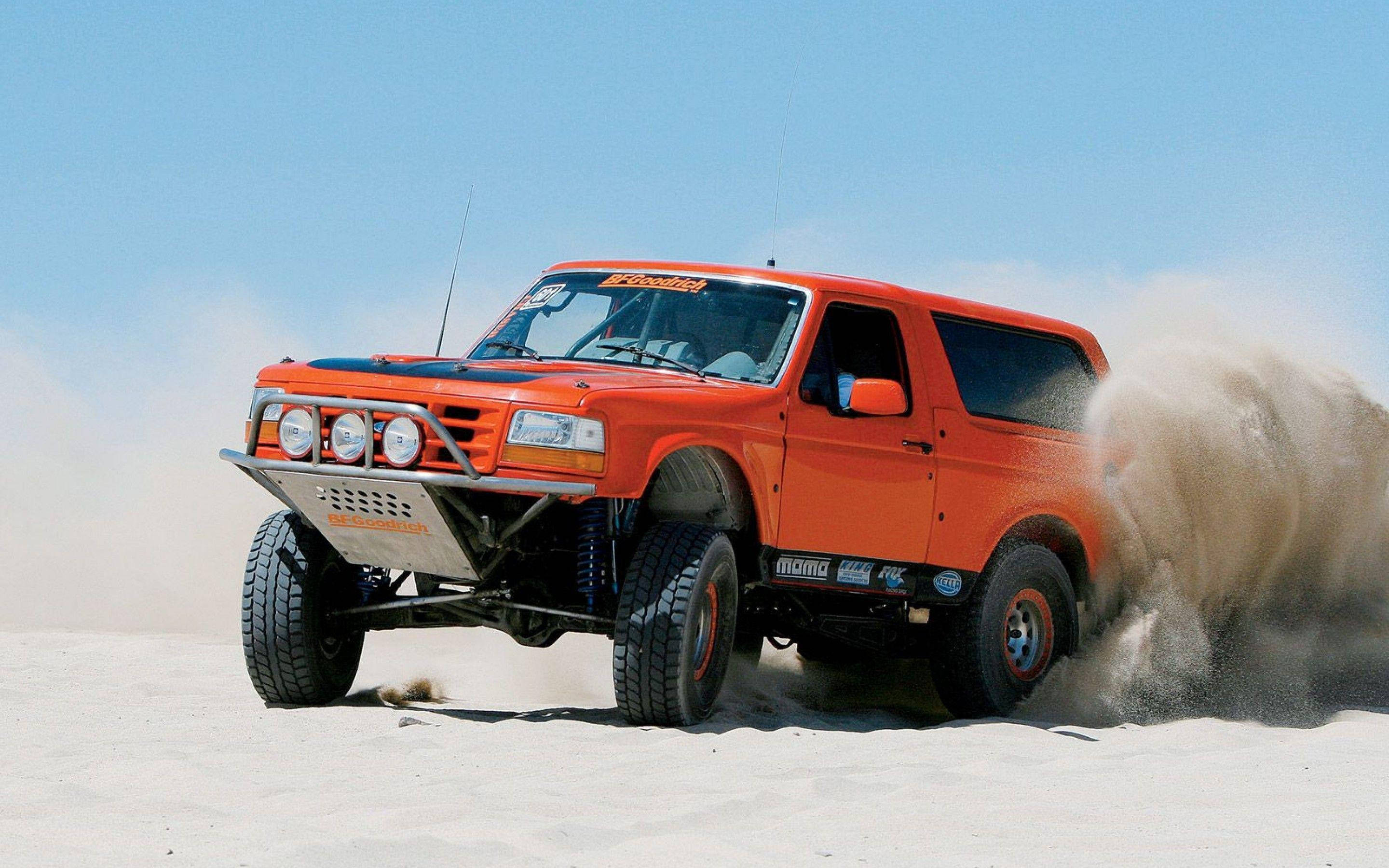 行駛於沙丘的福特 Bronco 壁紙 — 一輛亮橙色的越野卡車在晴朗的藍天下，高速穿越廣闊的沙漠，揚起沙塵。