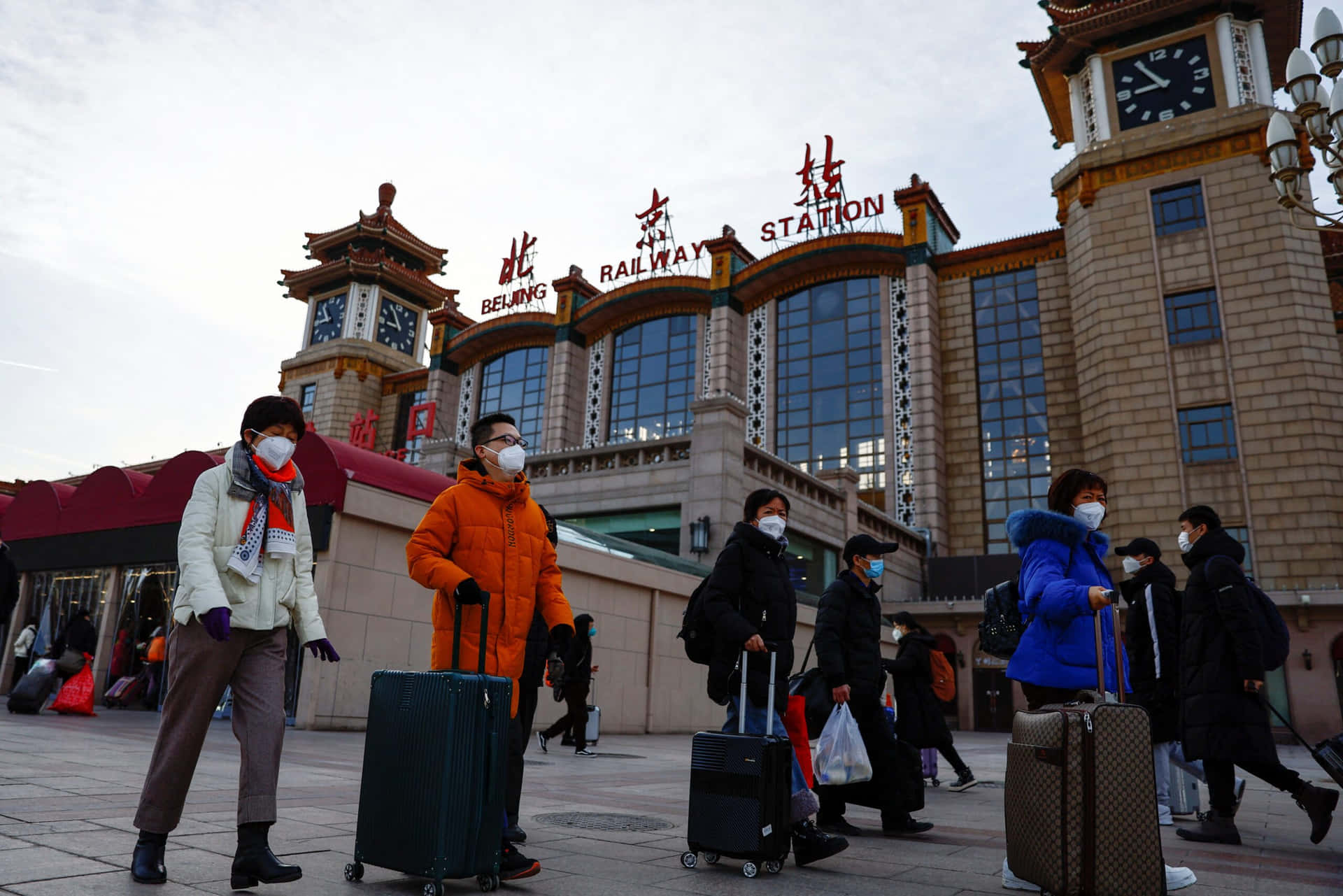 街燈照亮的中文字符 壁紙 — 旅客拖著行李箱、戴著口罩，在陰天下的北京火車站外行走。