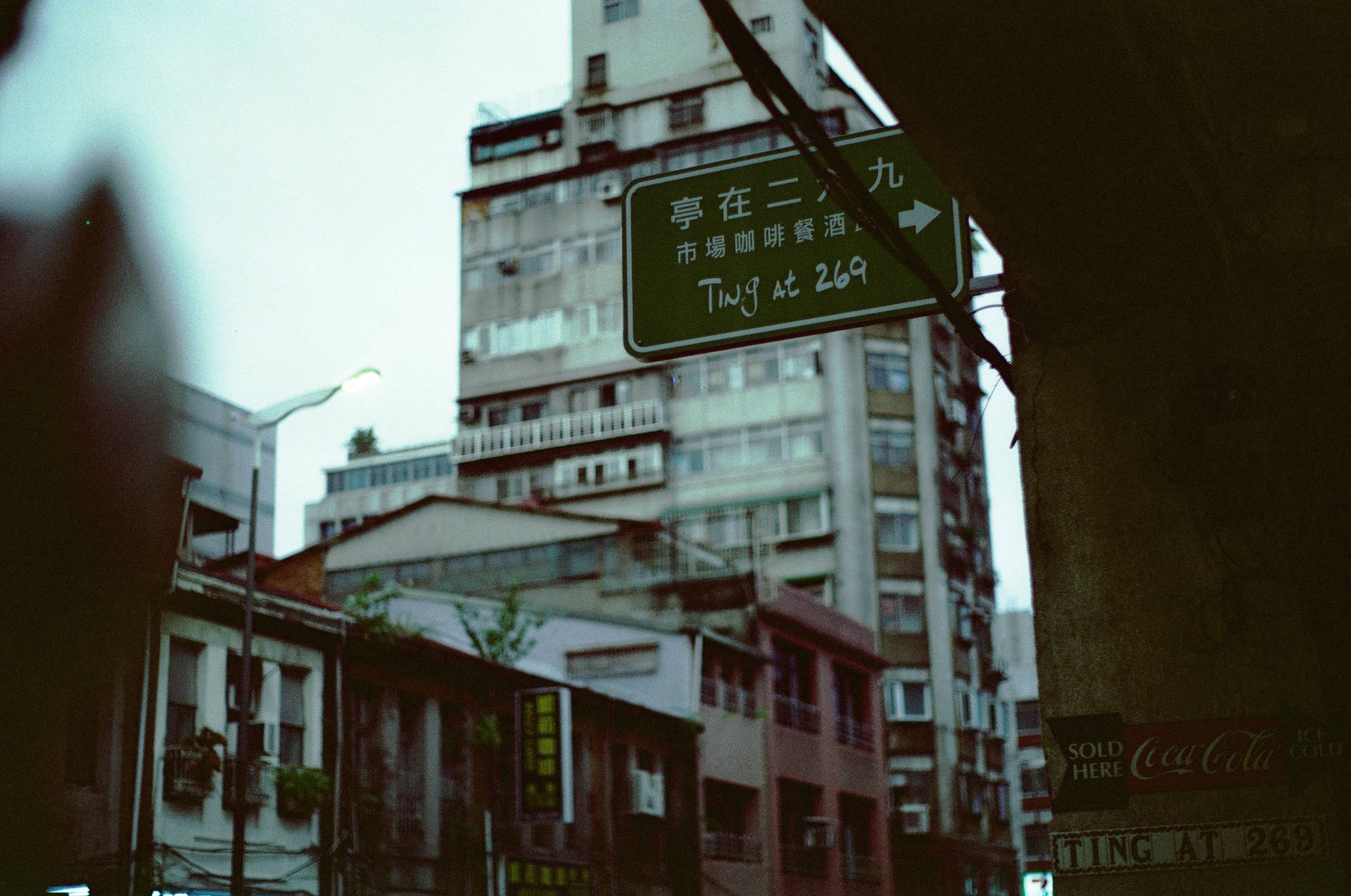 一塊褪色的綠色路牌，上面有手寫文字，掛在狹窄的城市小巷中。
