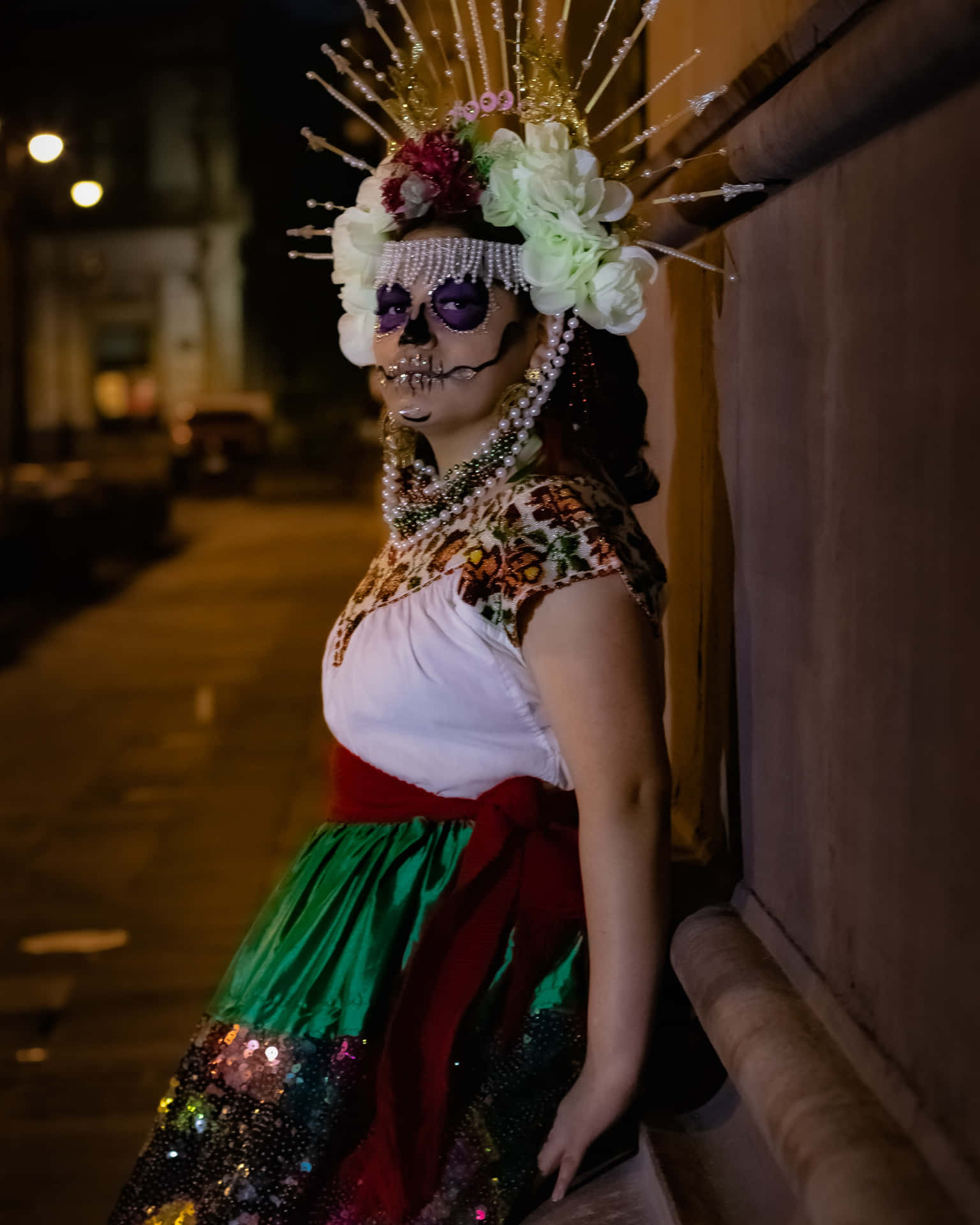 身穿色彩繽紛亡靈節服裝、化著骷髏妝容的女性，在夜晚靠著牆壁。 — 墨西哥 GIF 壁紙