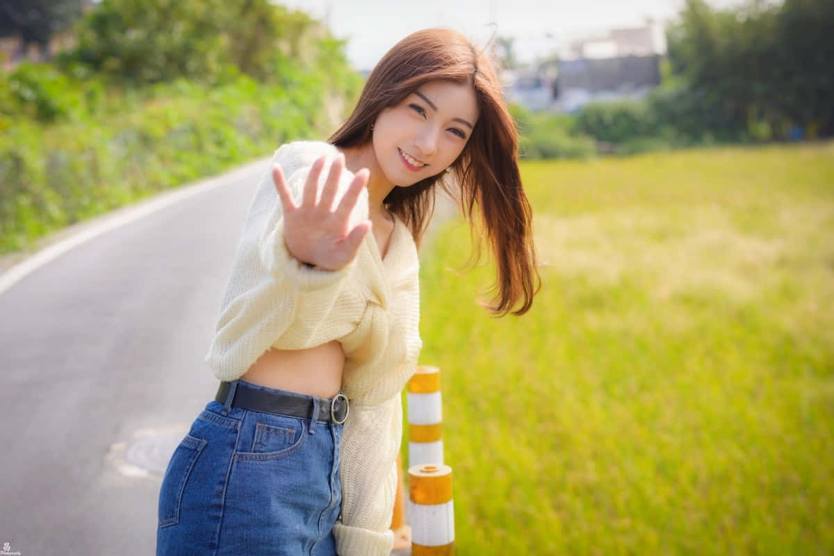 街頭微笑的亞洲女性 壁紙 — 一位微笑的年輕女子身穿米色毛衣和牛仔褲，在路邊護欄旁愉快地揮手，身後是茂密的綠草和蜿蜒的道路。