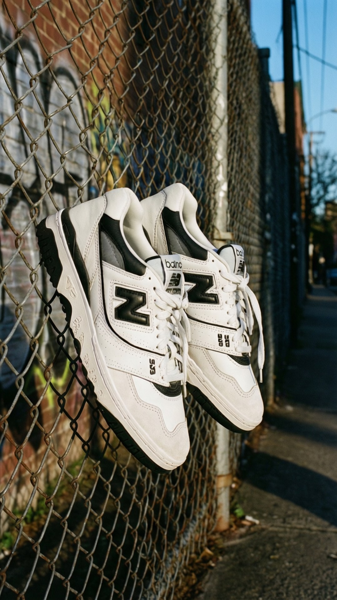 街頭懸掛的運動鞋 壁紙 — 一對帶有黑色點綴的白色 New Balance 運動鞋掛在鐵絲網圍欄上，背景是帶有塗鴉的磚牆。