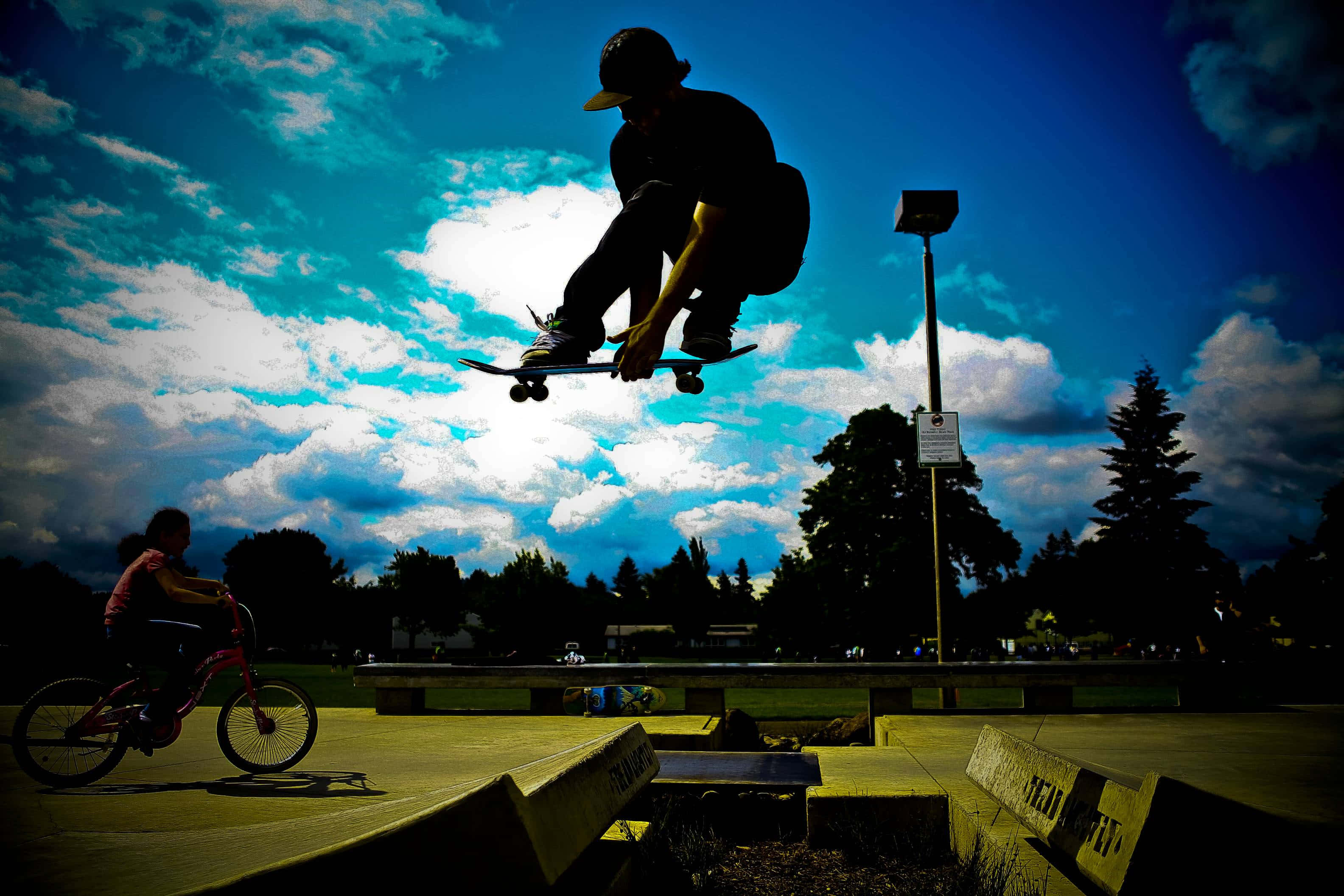 街頭滑板手表演 Kickflip 壁紙 — 一名滑板手在空中形成剪影，背景是充滿活力的藍天和蓬鬆的白雲，附近有一個小孩在騎單車。