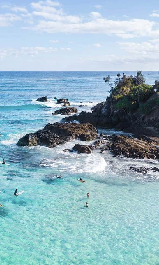 衝浪海灘美學圖片 壁紙 — 清澈的碧綠海水拍打著深色火山岩，衝浪者和游泳者在陽光明媚的海岸天堂中盡情享受。