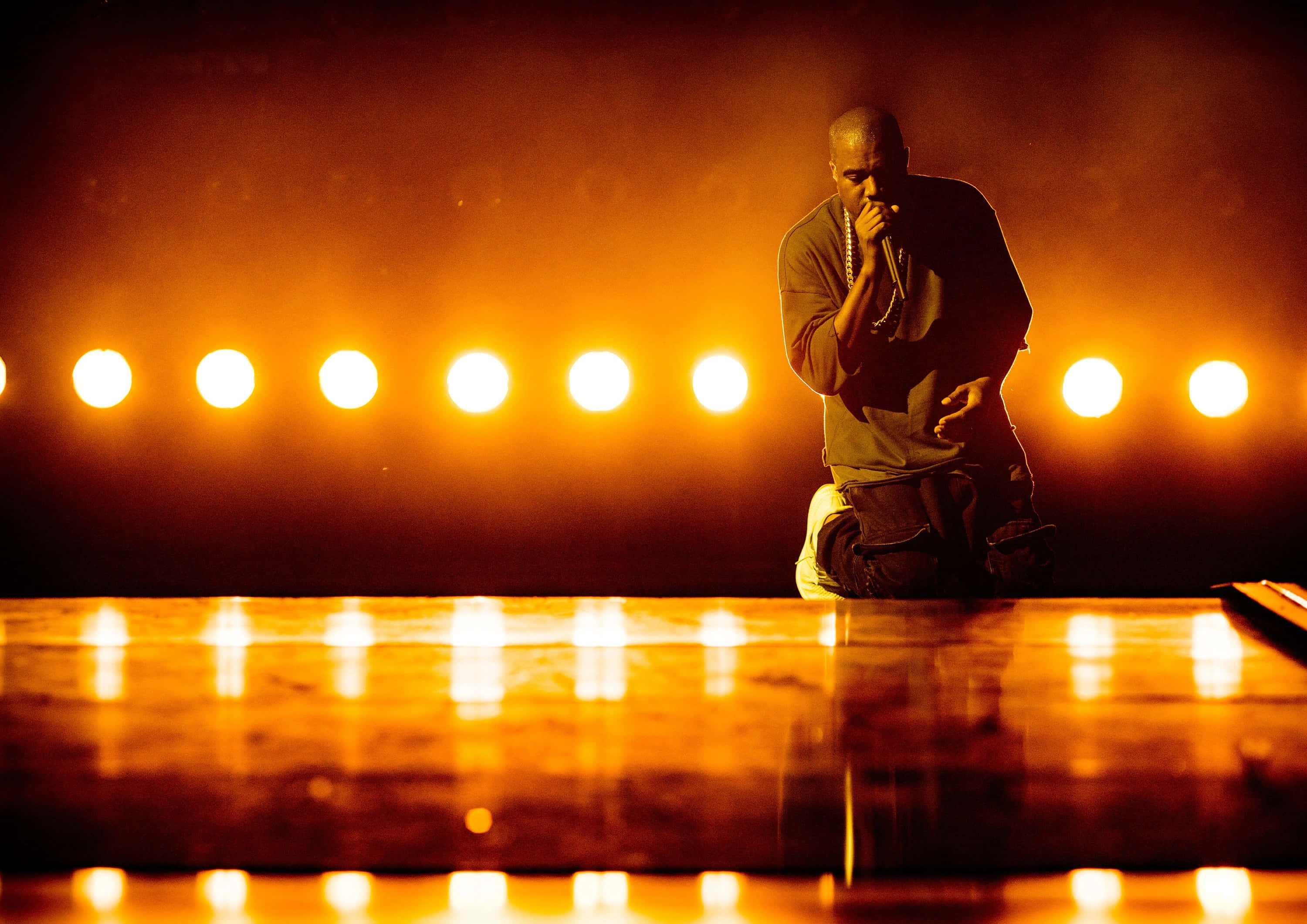 Kanye 桌布 壁紙 — 表演者在反射舞台上跪下，在溫暖的橙色燈光下，營造出戲劇性的音樂會時刻。