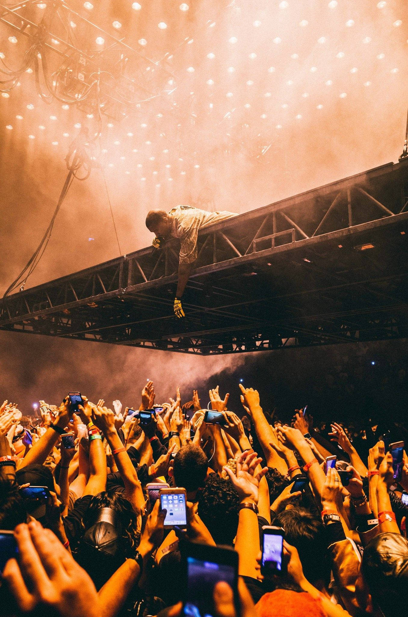 Kanye West Stage Presence 壁紙 — 表演者懸掛在舞台桁架上，下方是舉起手機的人海，捕捉著電音演唱會的能量。